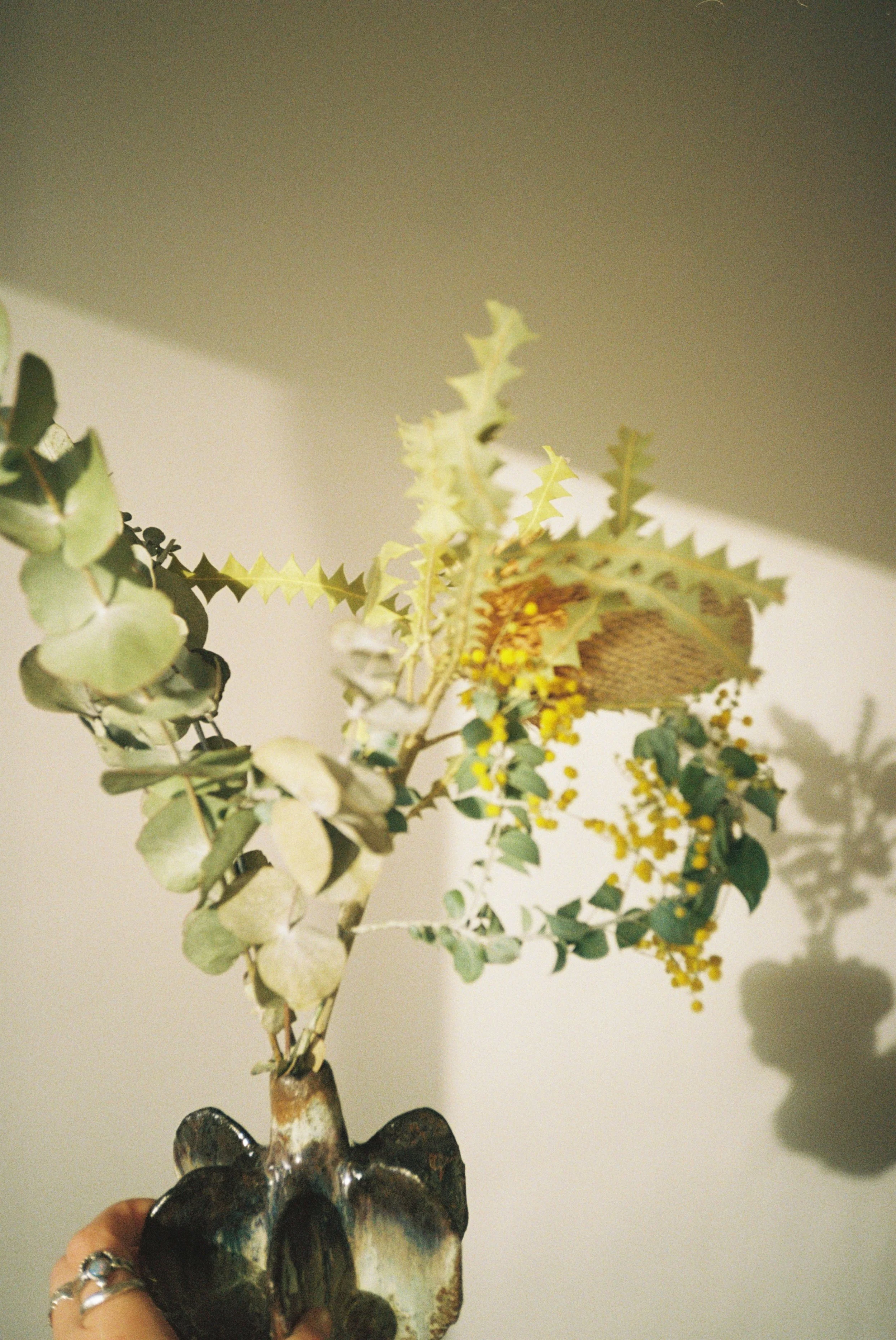 A small floral arrangement with green leaves, yellow flowers, and a decorative vase, against a neutral background with shadows.