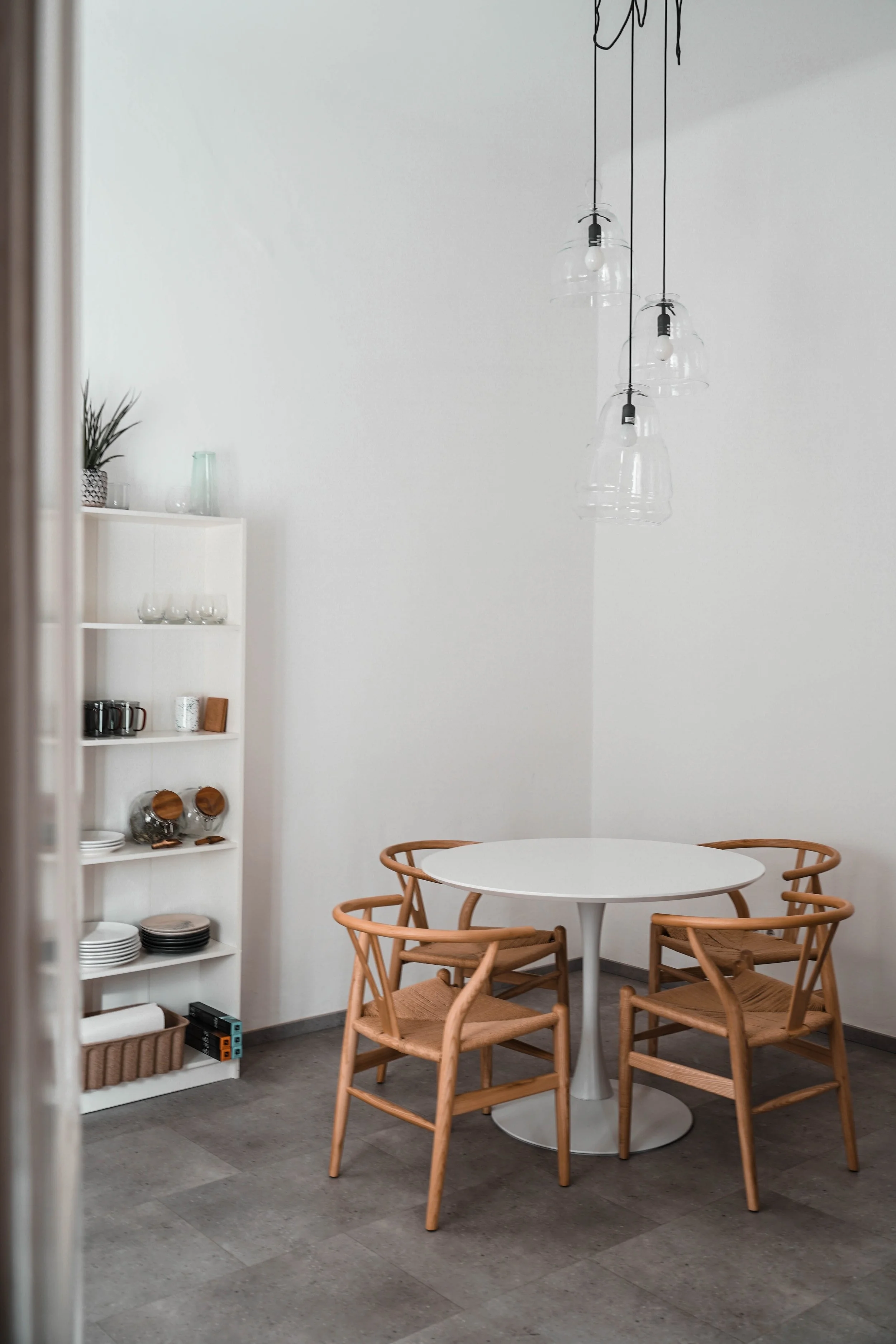 A minimalist dining area with a white round table, four wooden chairs with woven seats, a white shelving unit with dishes and glasses, and a modern light fixture with multiple hanging glass lamps.