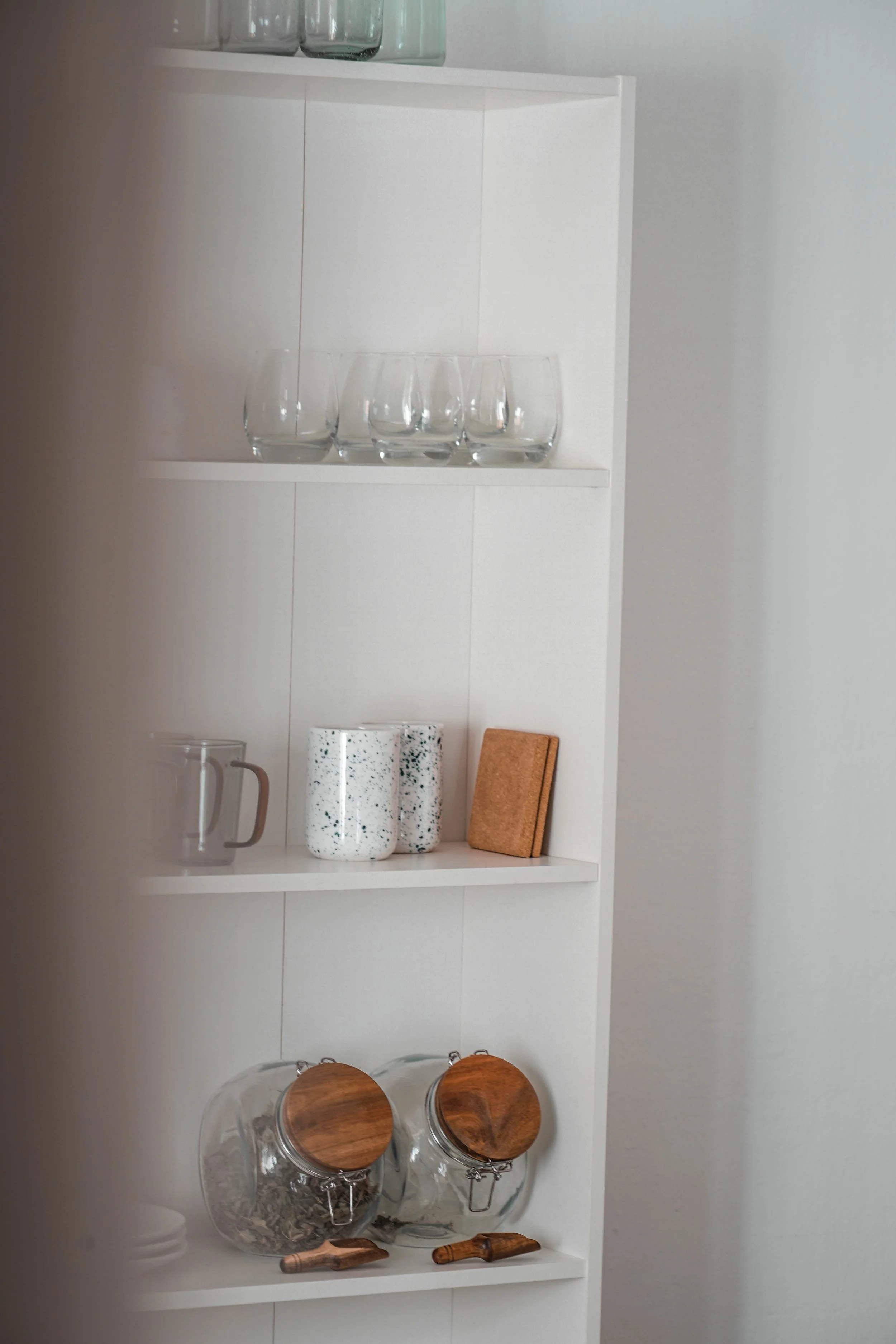 White kitchen shelf with clear glasses, speckled mugs, a small book, and glass jars with wooden lids.