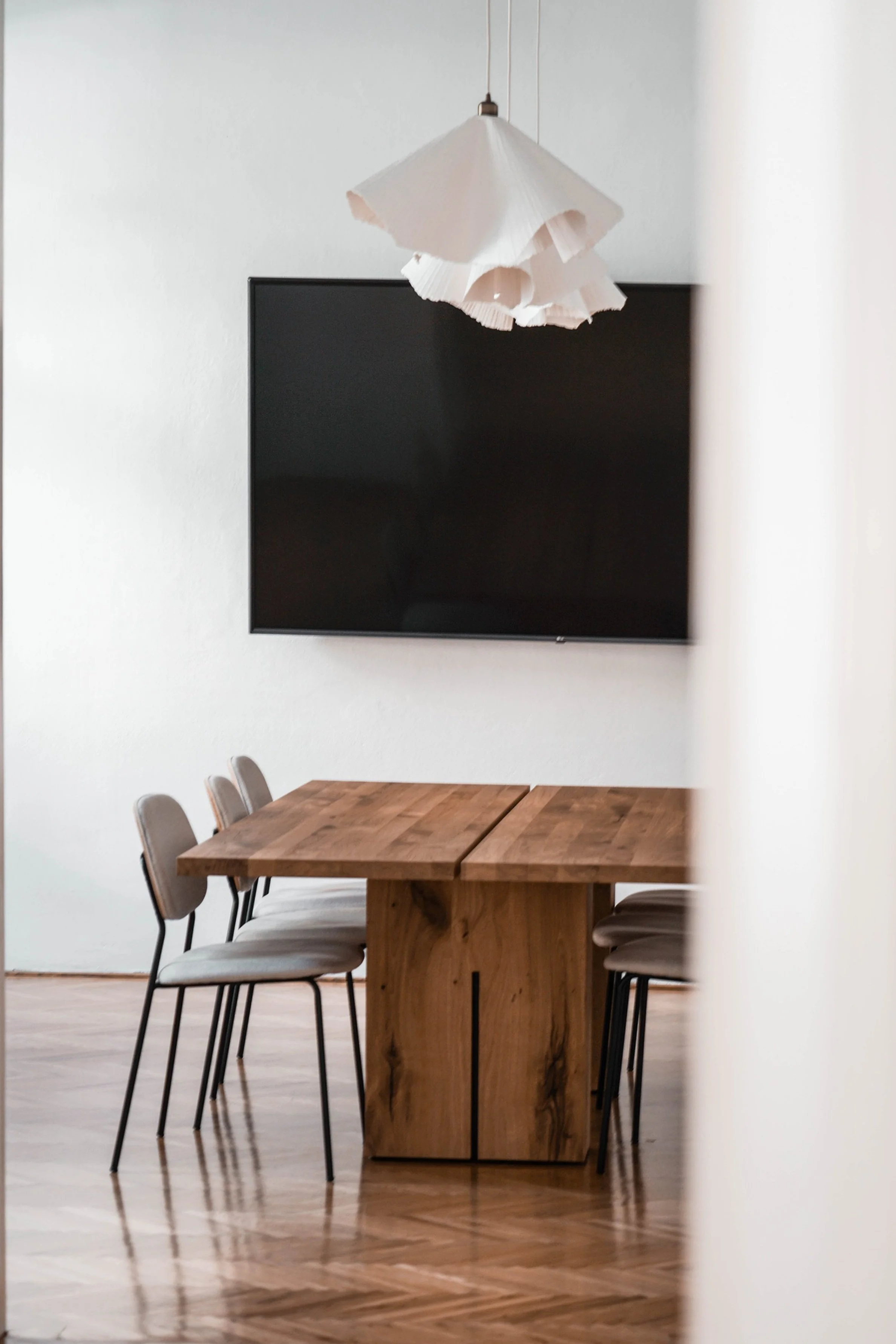 A dining area with a wooden table and six beige chairs, a large wall-mounted black television, and a white pendant light hanging from the ceiling.