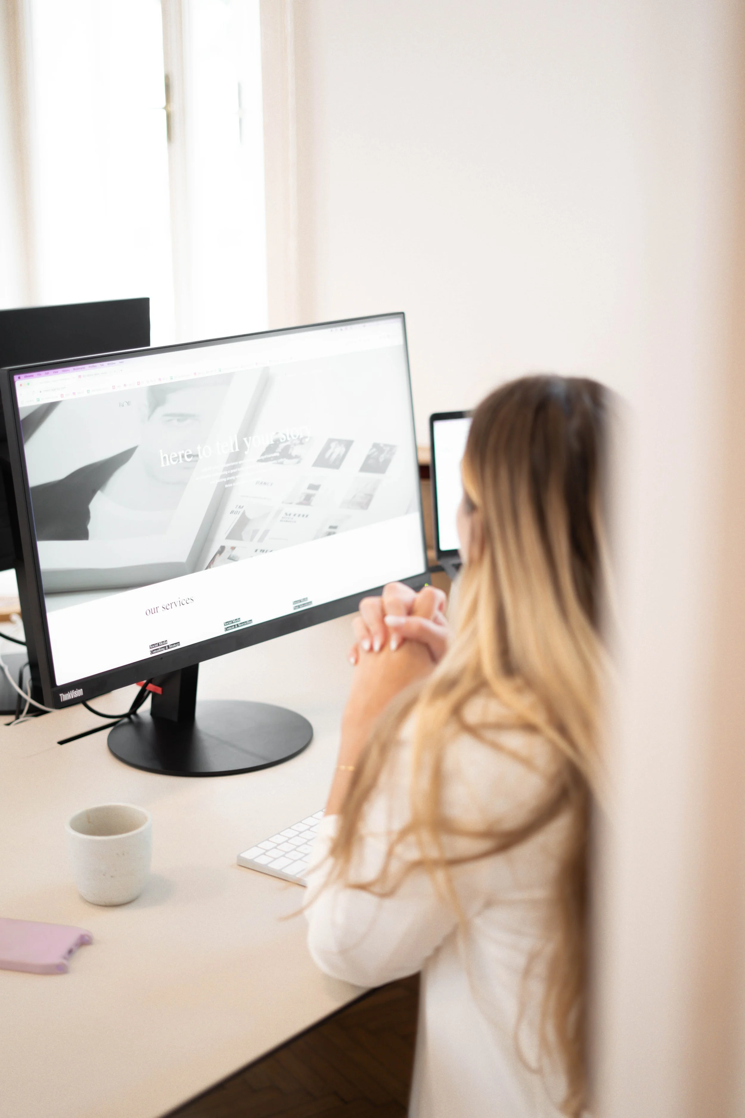 A woman with long blond hair sitting at a desk, looking at a computer monitor displaying a website.