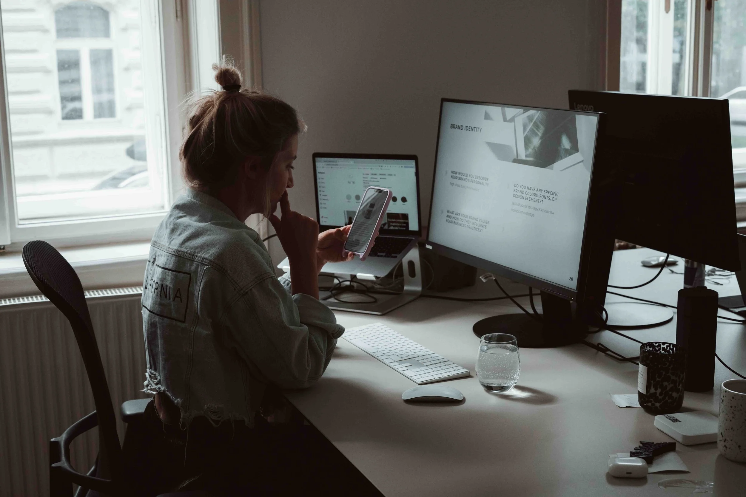 A woman sitting at a desk, looking at her phone, with two computer monitors, a keyboard, mouse, and various items on the desk, near a window with natural light.