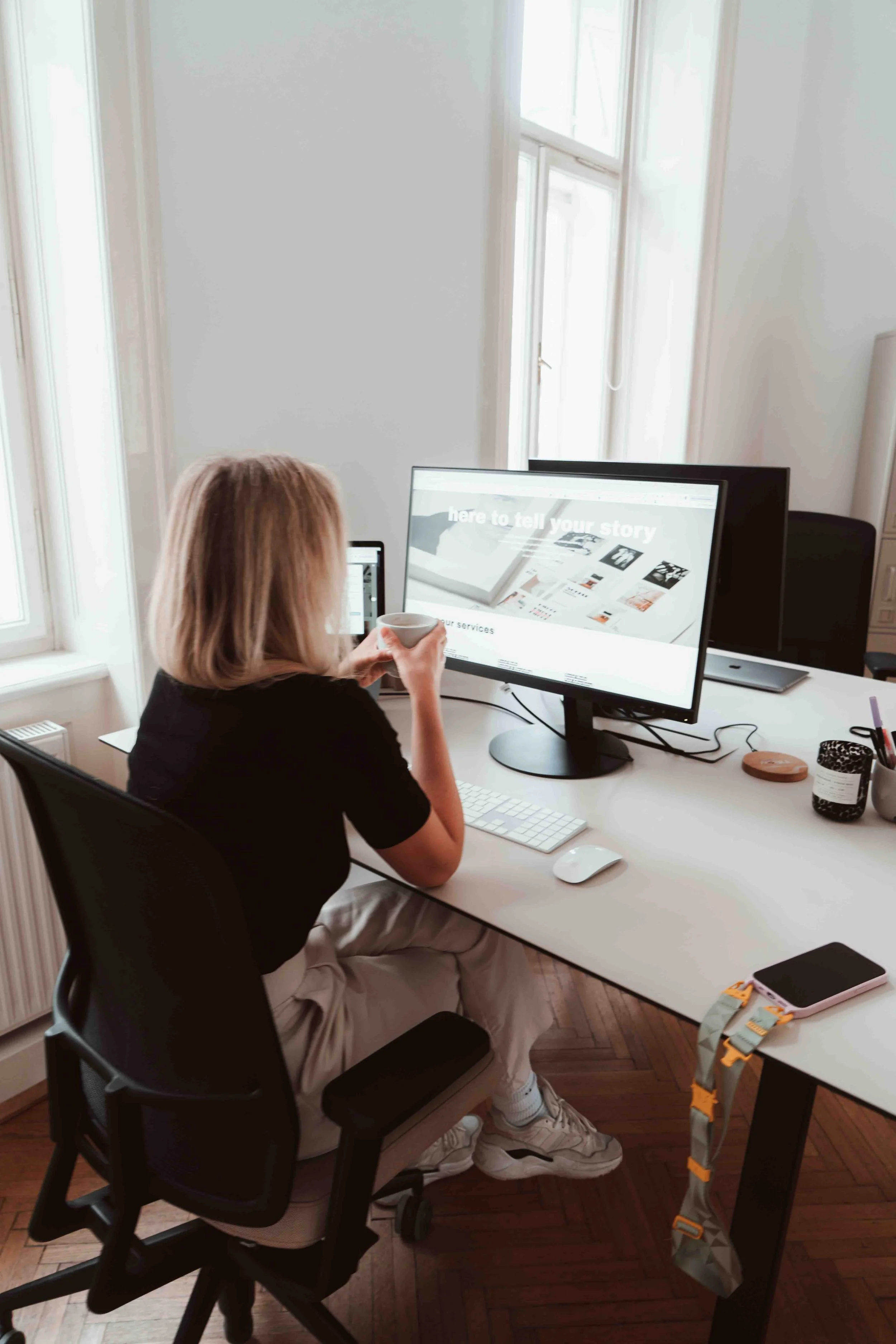 A woman sitting at a desk, drinking from a cup, working on a large computer monitor in a bright room with large windows.