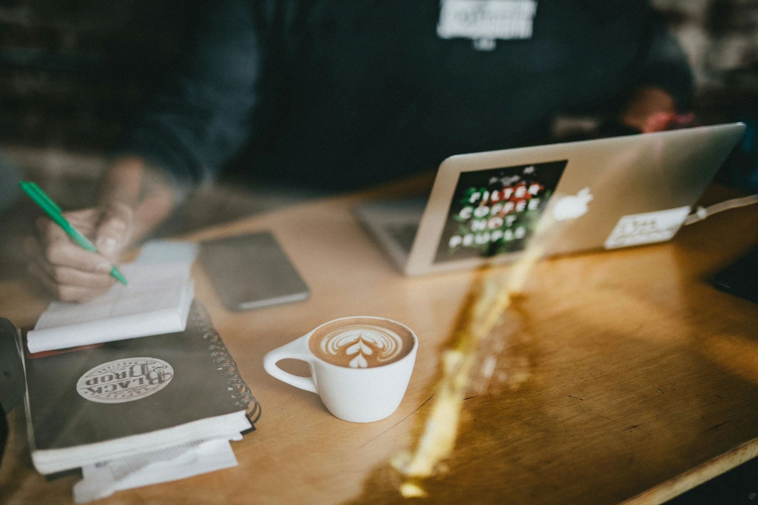 person working at a coffee shop writing on a notepad surrounded by laptop, phone and cup of coffee