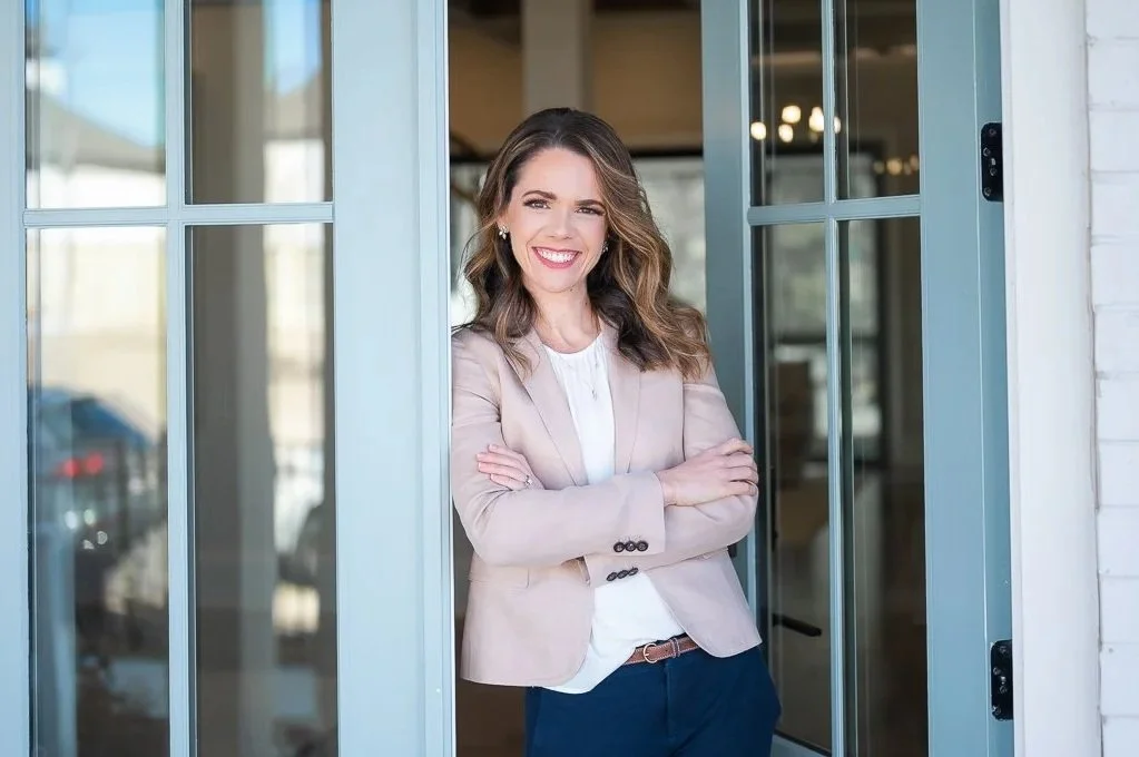female real estate agent in a beige blazer leaning against an open light blue door at a luxury real estate listing