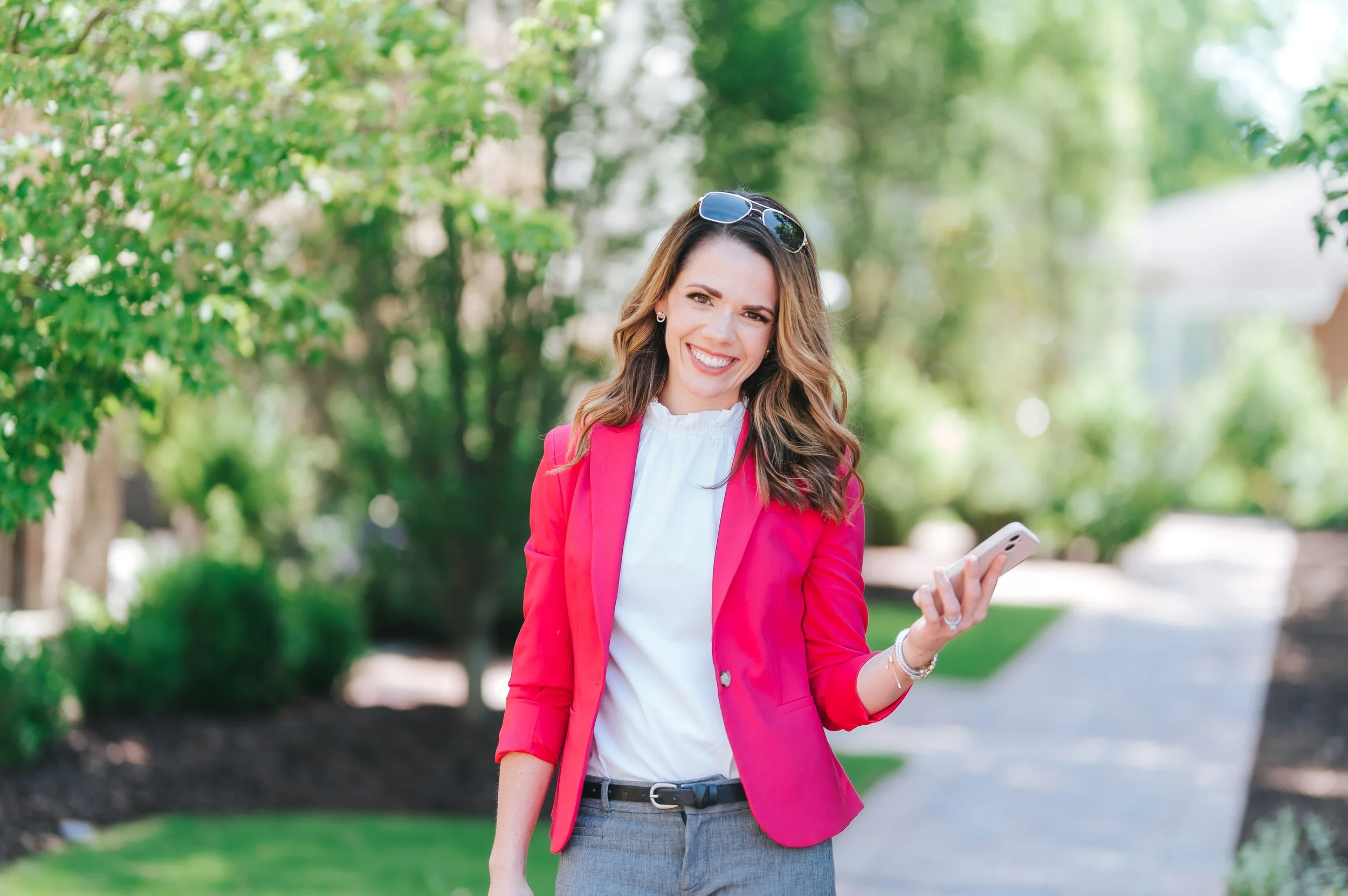 female real estate agent with bright pink jacket walking in green space in Alpharetta, Georgia while holding a light pink iphone