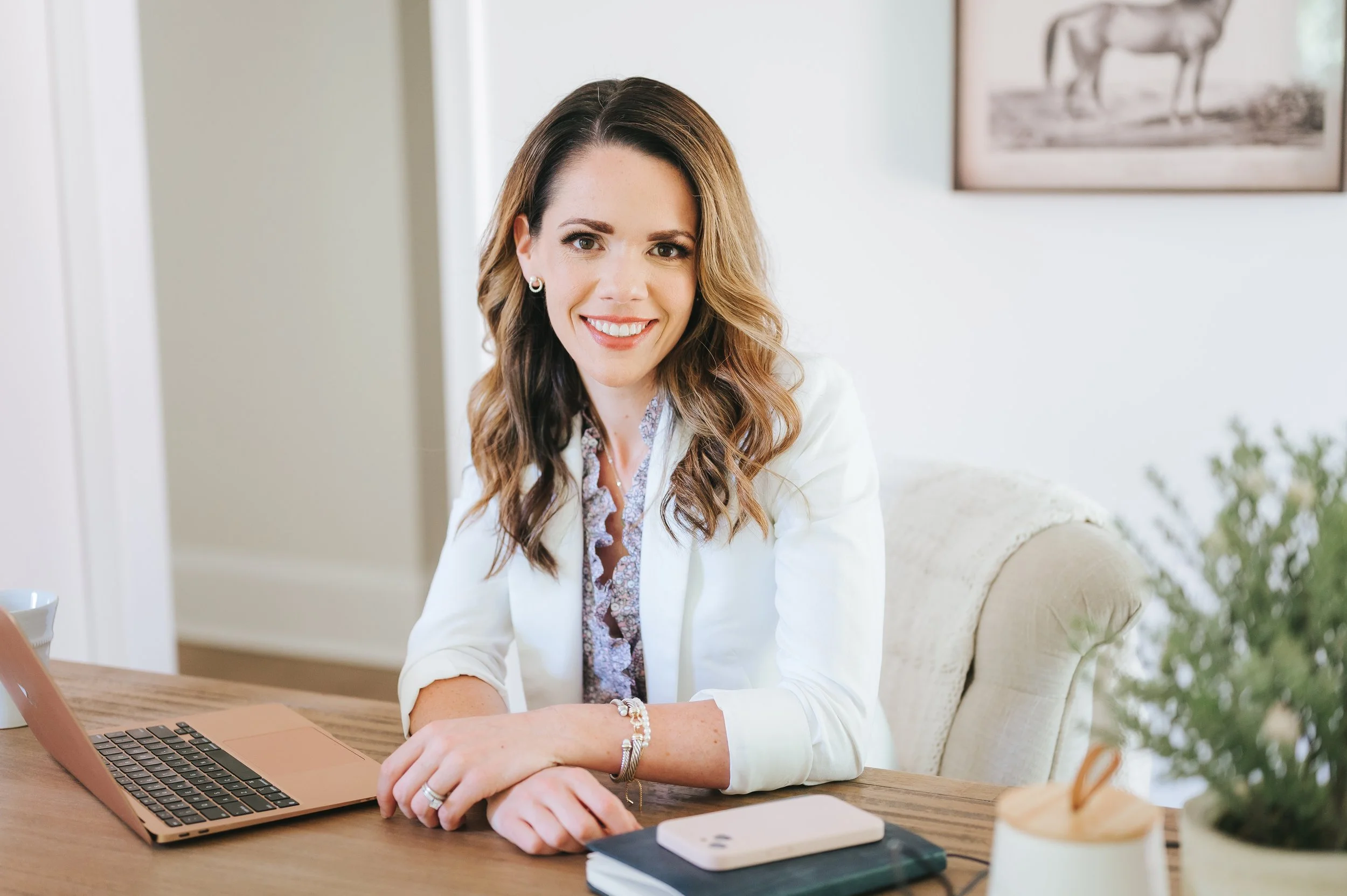 female real estate agent in white blazer sitting at a wooden desk working at her laptop