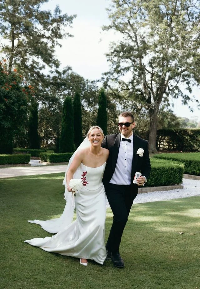 A bride and groom walking together outdoors on a sunny day, smiling and happy. The bride is wearing a white wedding dress with floral embroidery and holding a bouquet, while the groom is dressed in a black tuxedo with sunglasses and holding a drink.