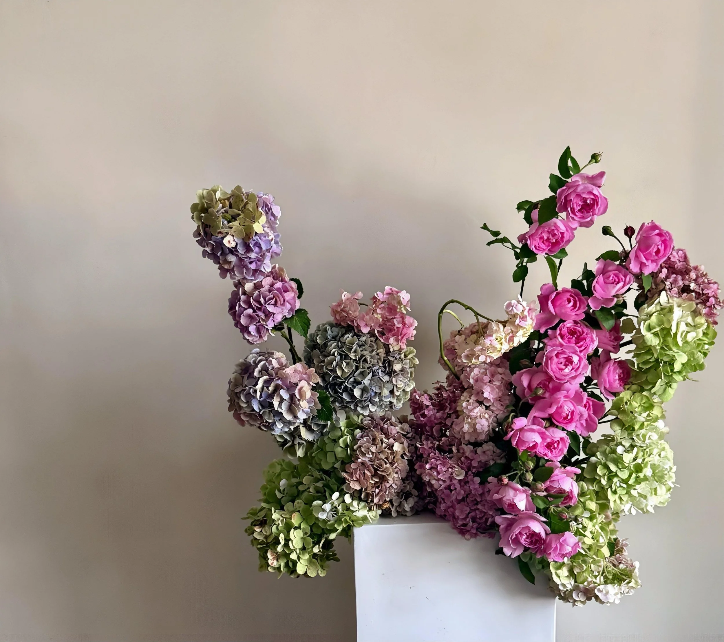 Arrangement of pink and purple hydrangea flowers on a white stand with a plain, light-colored wall background.