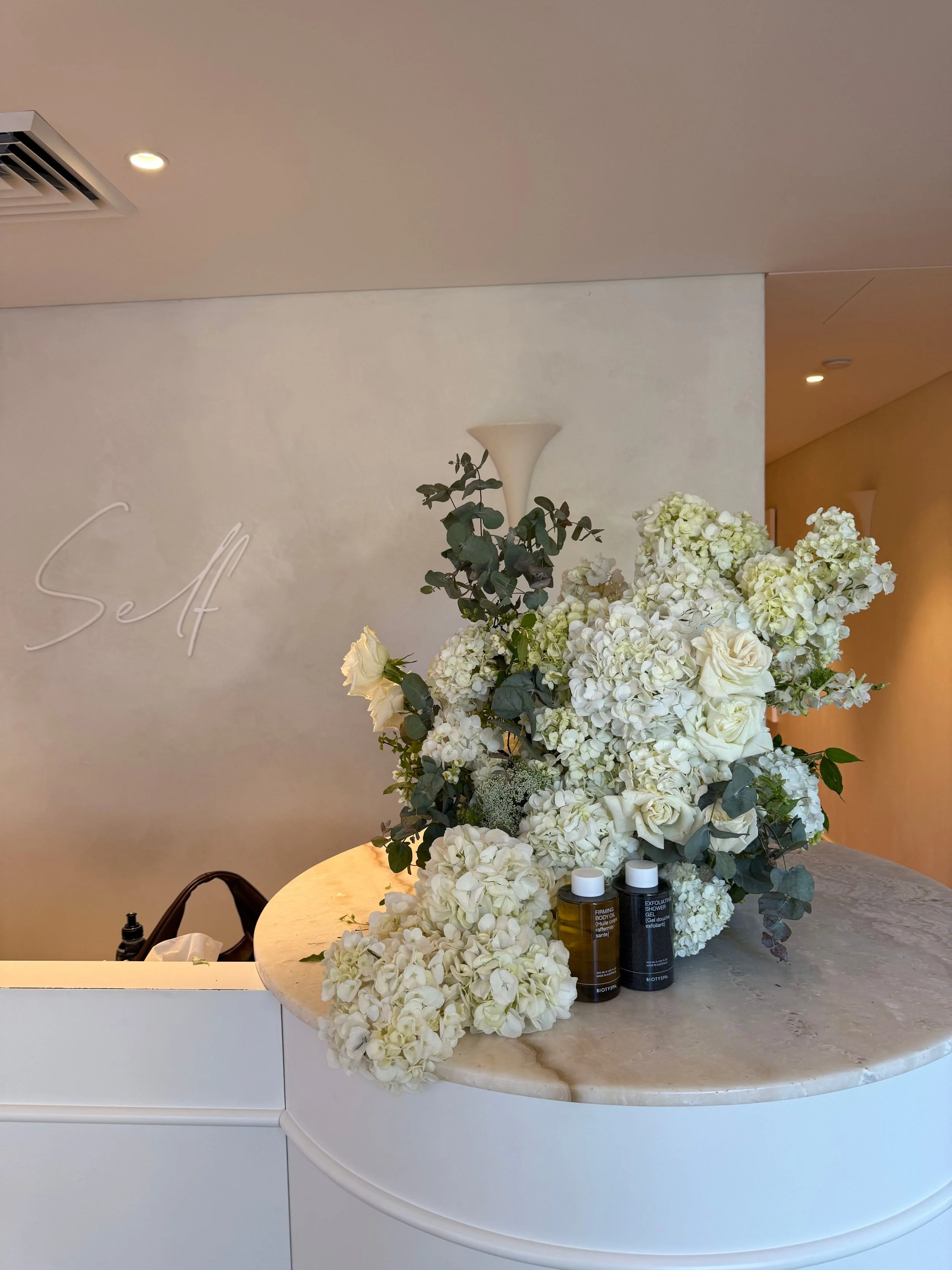 Elegant floral arrangement of white flowers and greenery on a marble-topped counter with skincare bottles. Background features a beige wall with decorative lighting and a minimalist wall sconce.
