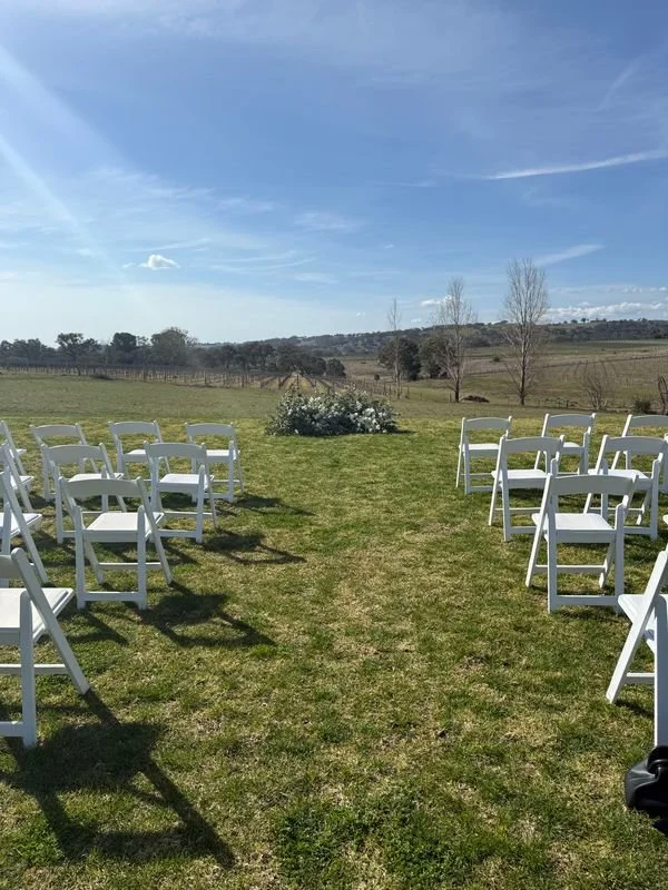 Outdoor wedding ceremony setup with white chairs arranged on a grassy field, facing an altar of flowers with a rural landscape and blue sky in the background.