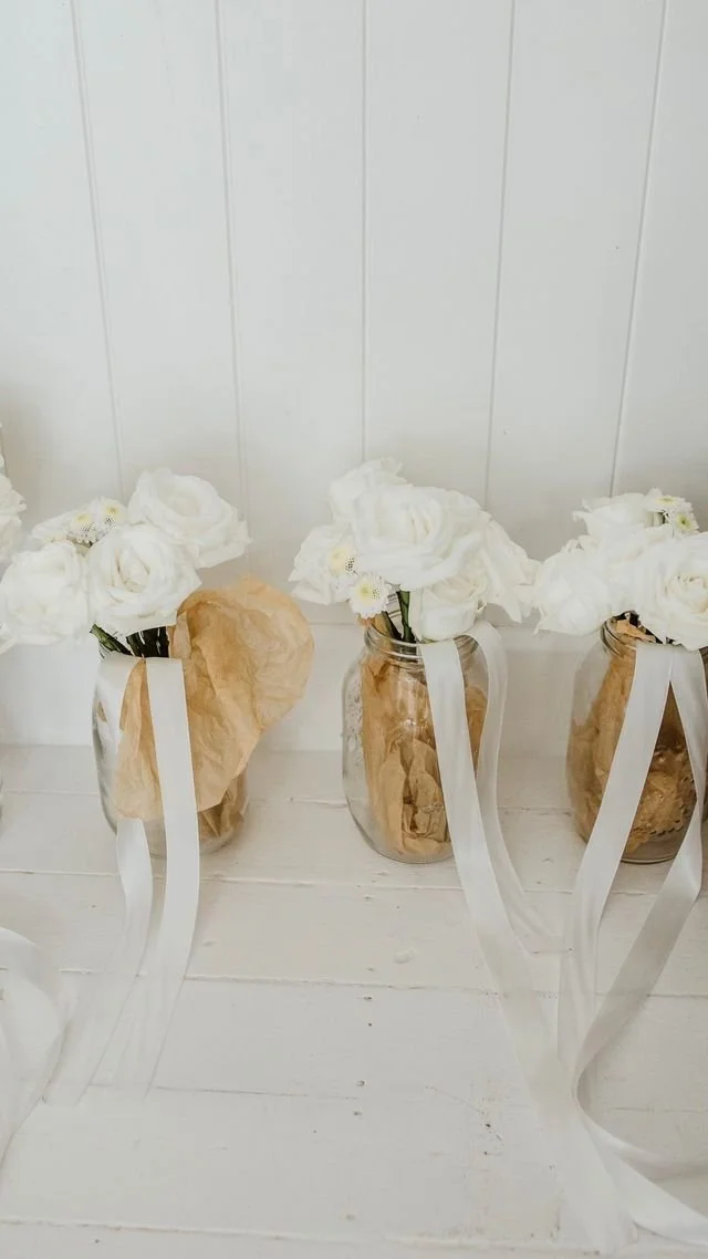 Three vases with white roses and small white flowers, wrapped in brown paper, with white ribbons attached.