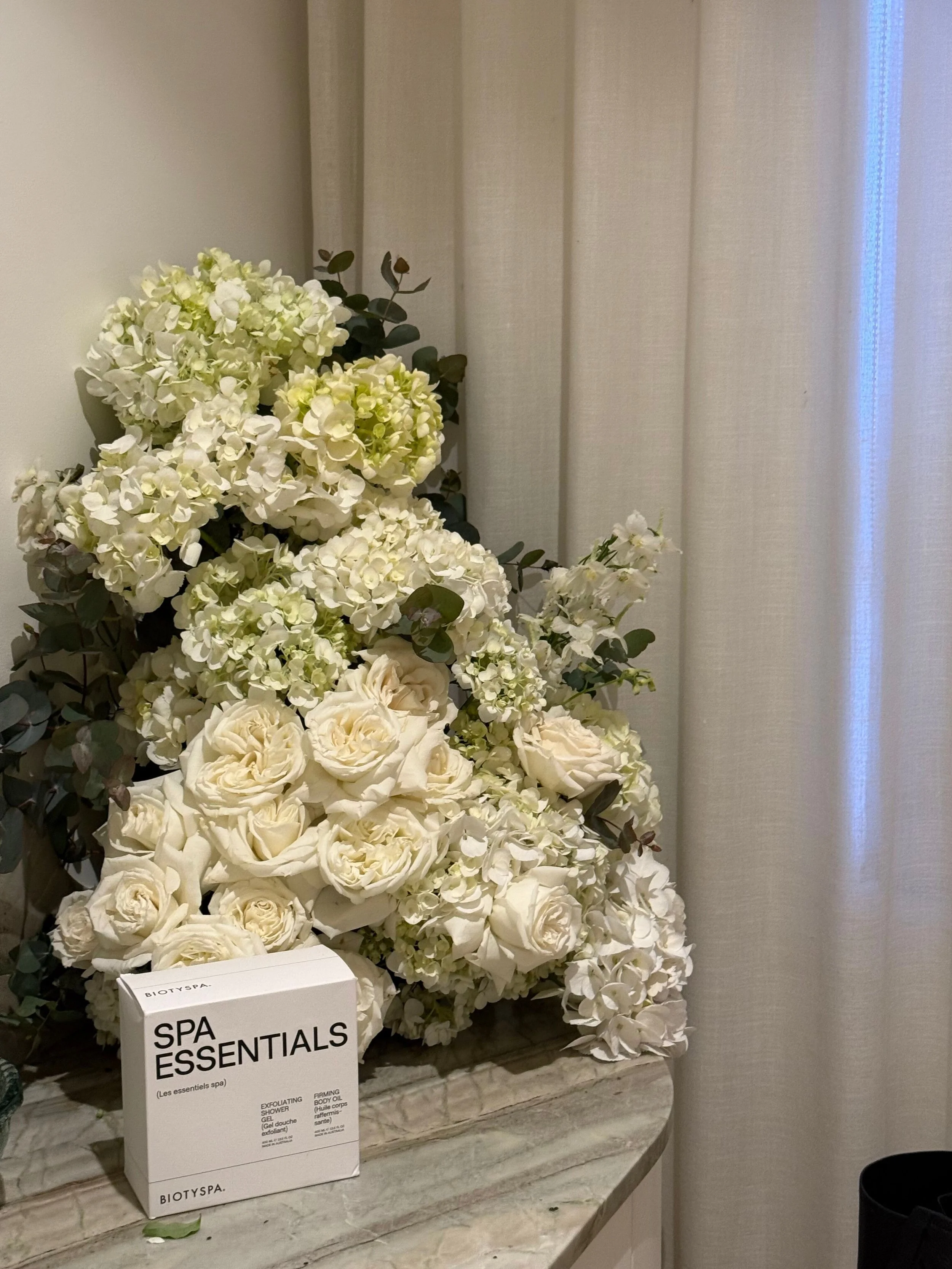White hydrangea and rose flower arrangement on a marble countertop, with a box of Biotyspa SPA Essentials in front.