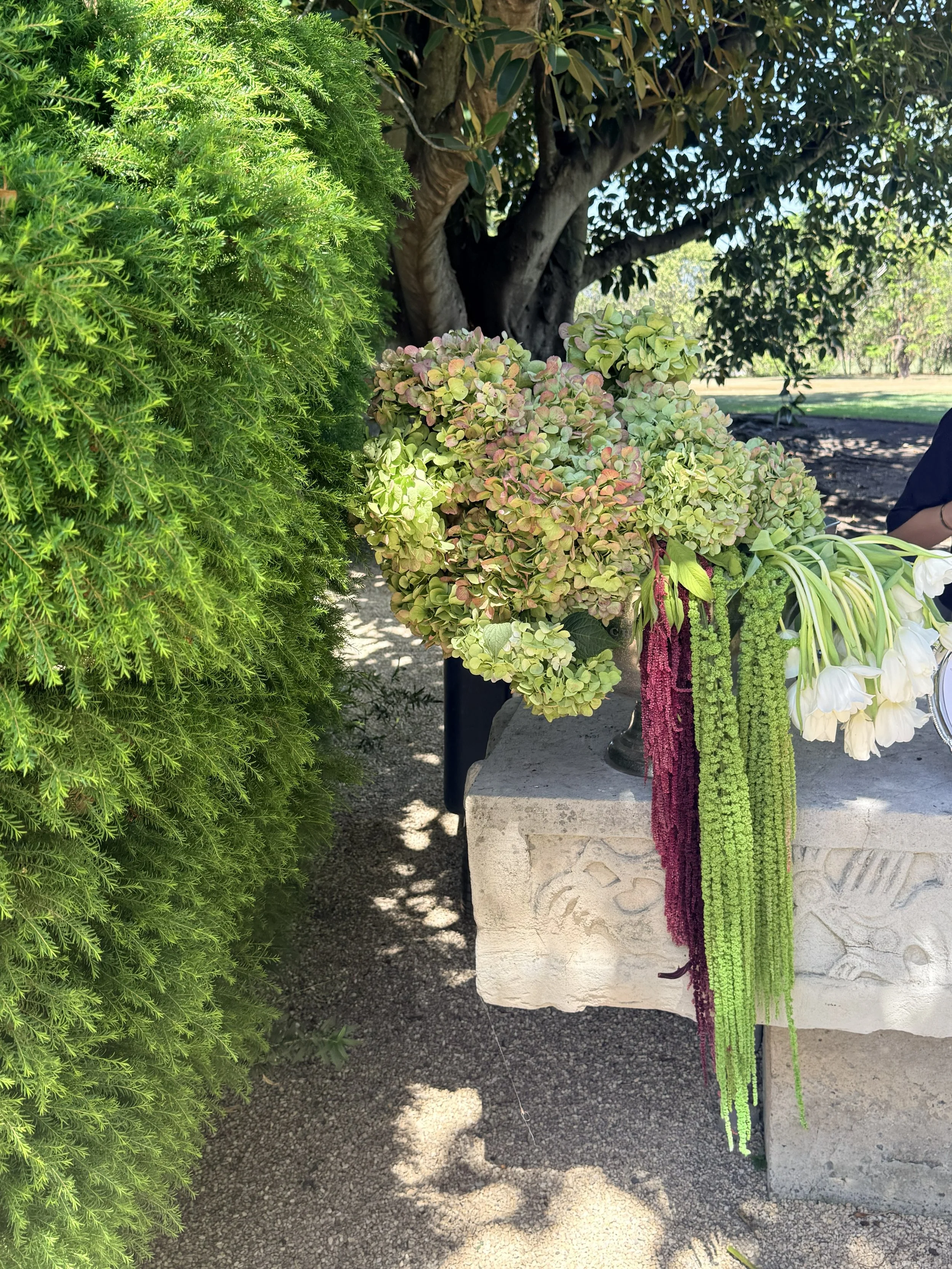 Colorful floral arrangement on a stone pedestal outdoors, with lush green trees and a park setting in the background.