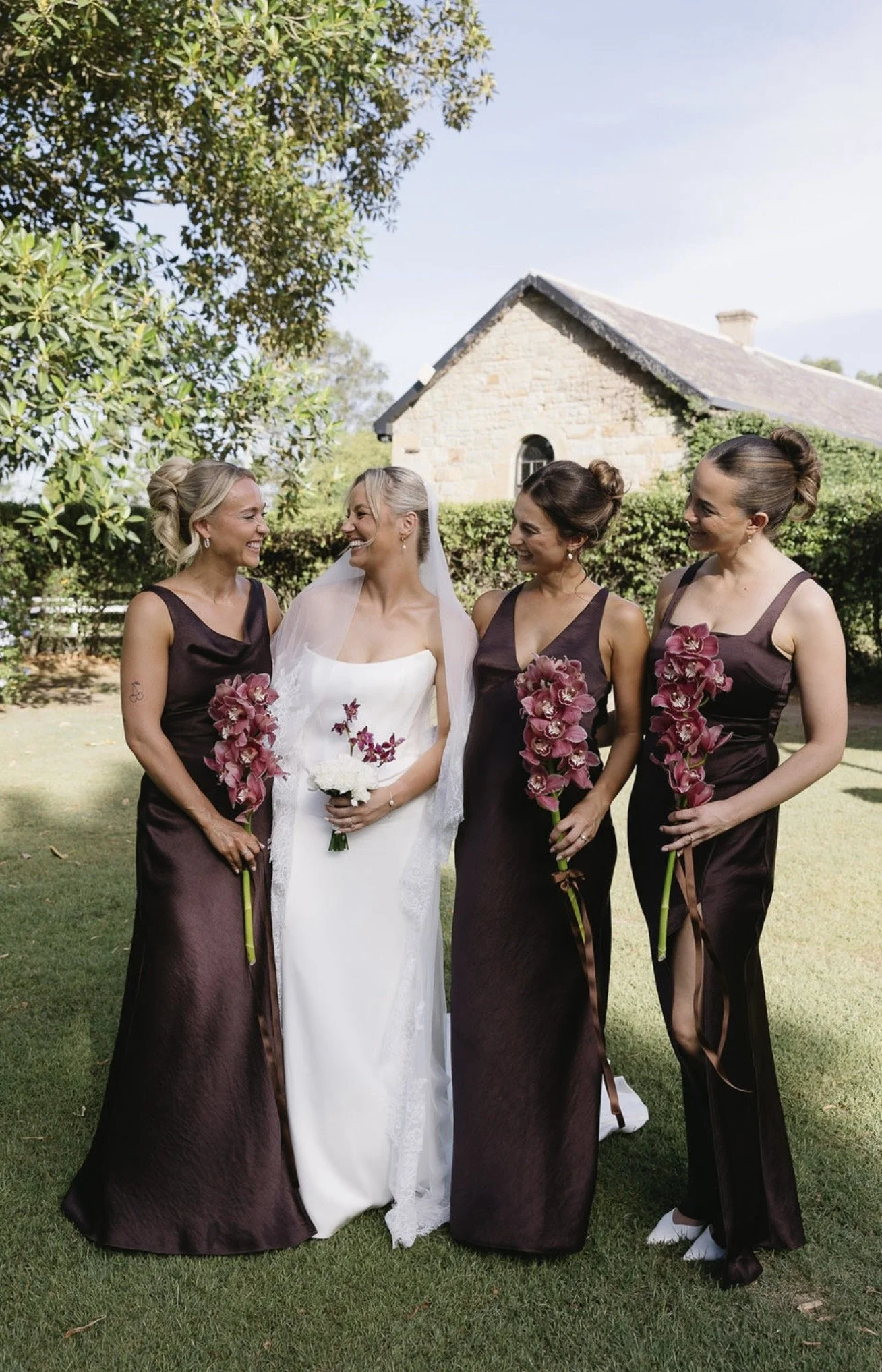 Four women standing outdoors on green grass near a building with a stone facade. One woman is wearing a white wedding dress with a veil and holding a bouquet, and the three women are dressed in matching dark brown dresses and holding pink flowers.