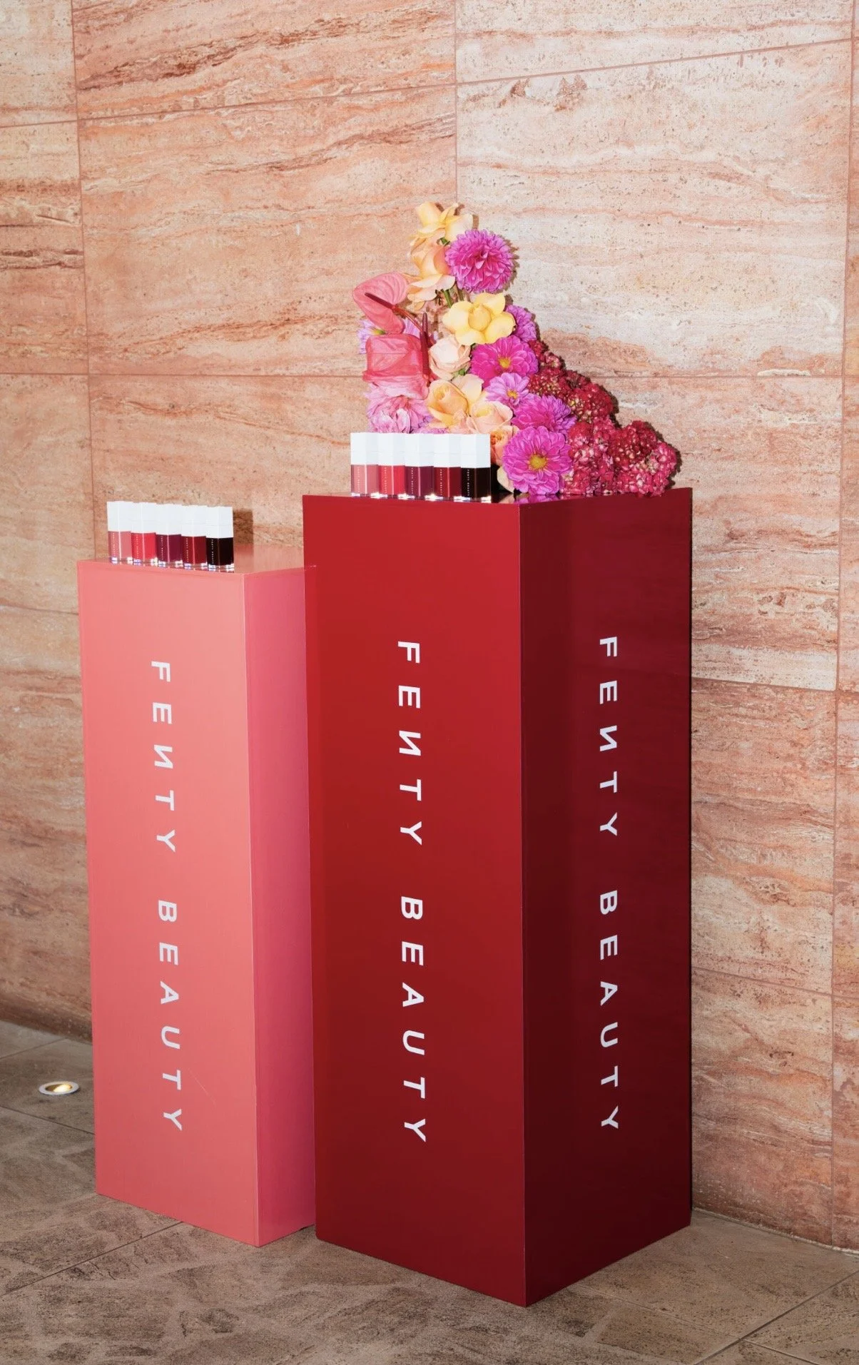 Two tall display stands with lipstick samples and flower arrangements in a lobby or showroom. The stands are pink and red, with the words "FENTY BEAUTY" written vertically.
