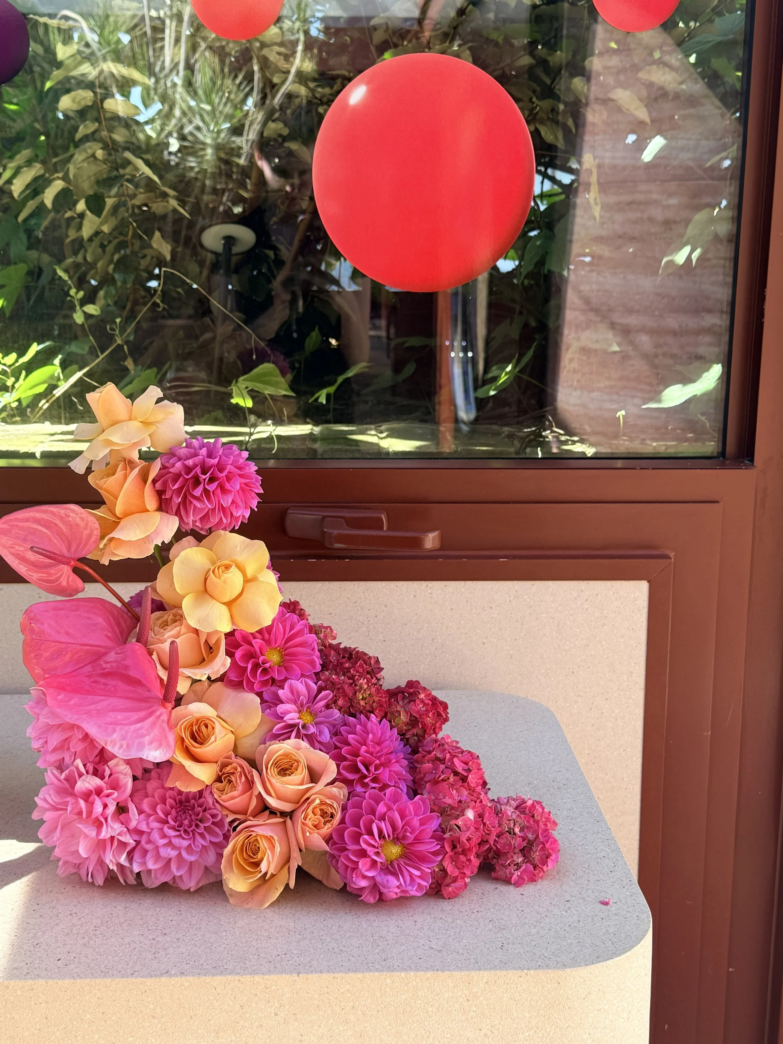 Colorful floral arrangement with pink dahlias, peach roses, pink anthuriums, and red hydrangeas on a light-colored surface outside a window with a red balloon and greenery visible in the background.