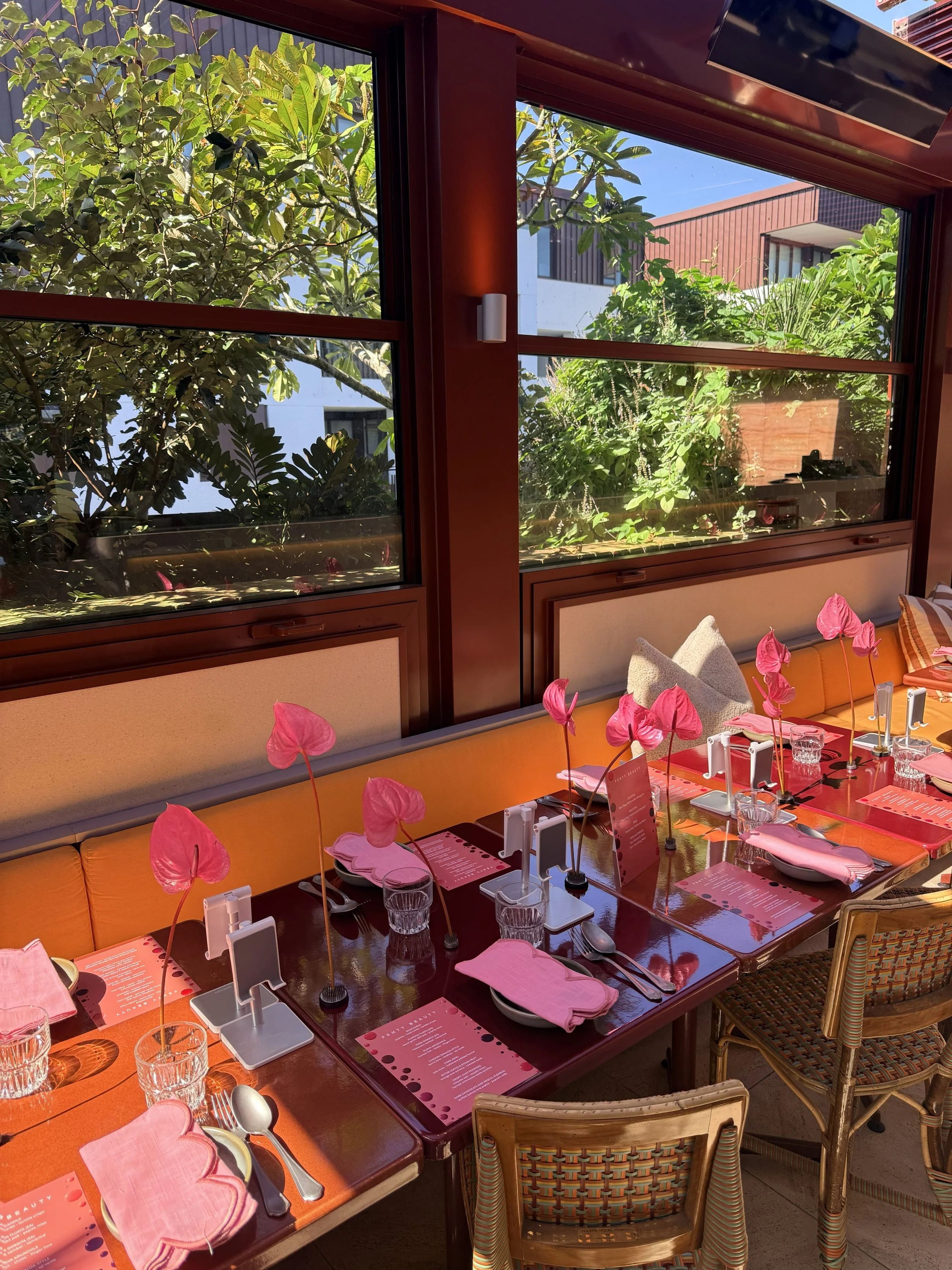 A cozy restaurant or cafe with a window view of lush green trees and modern buildings outside. Inside, a table is set with pink napkins shaped like hearts, glasses, utensils, pink menus, and heart-shaped pink decorations, creating a romantic or elega