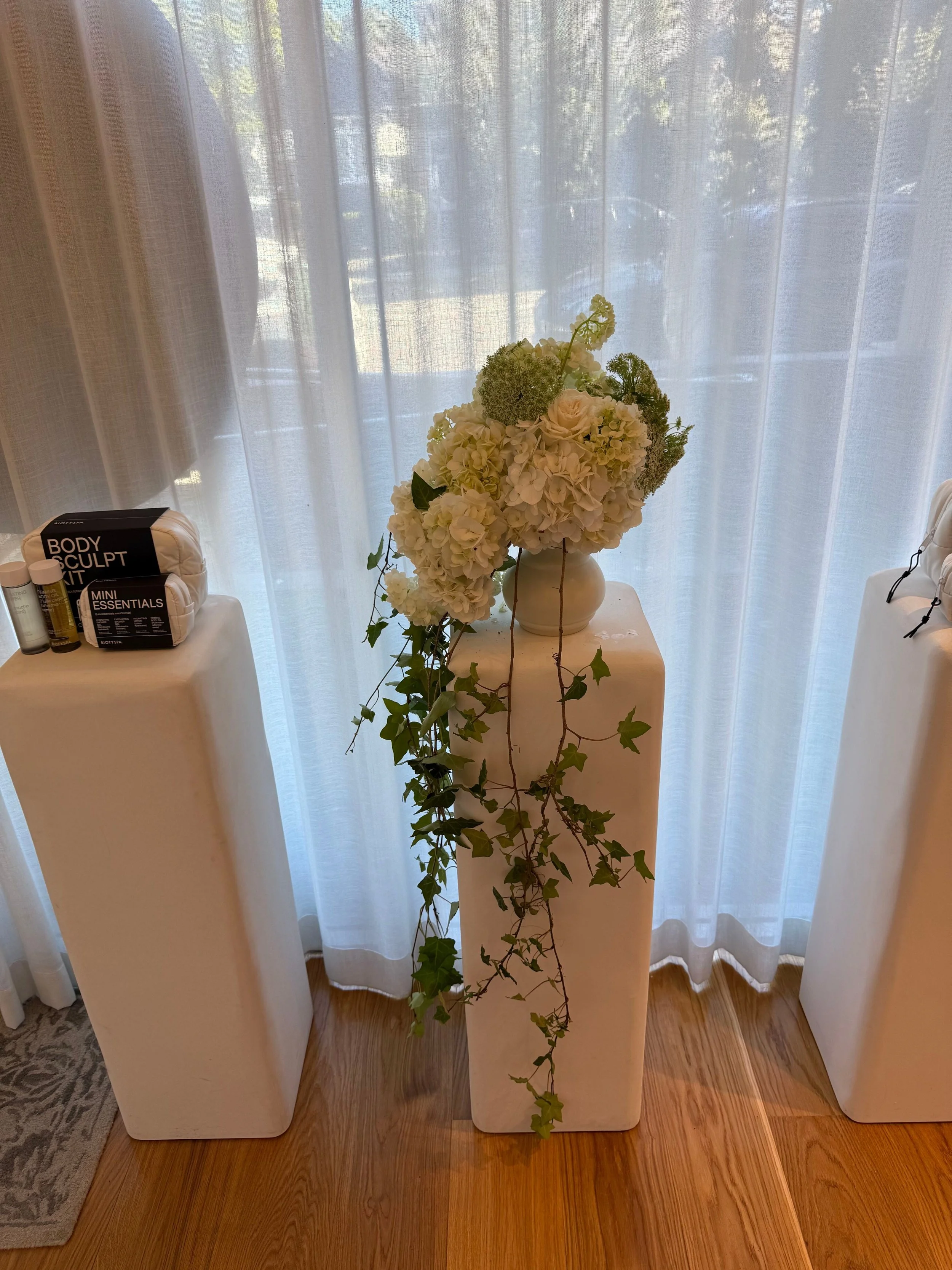 White rectangular pedestals with a floral arrangement of white and green flowers in a white vase, and green ivy spilling down the front pedestal, placed in front of sheer curtains covering a window.