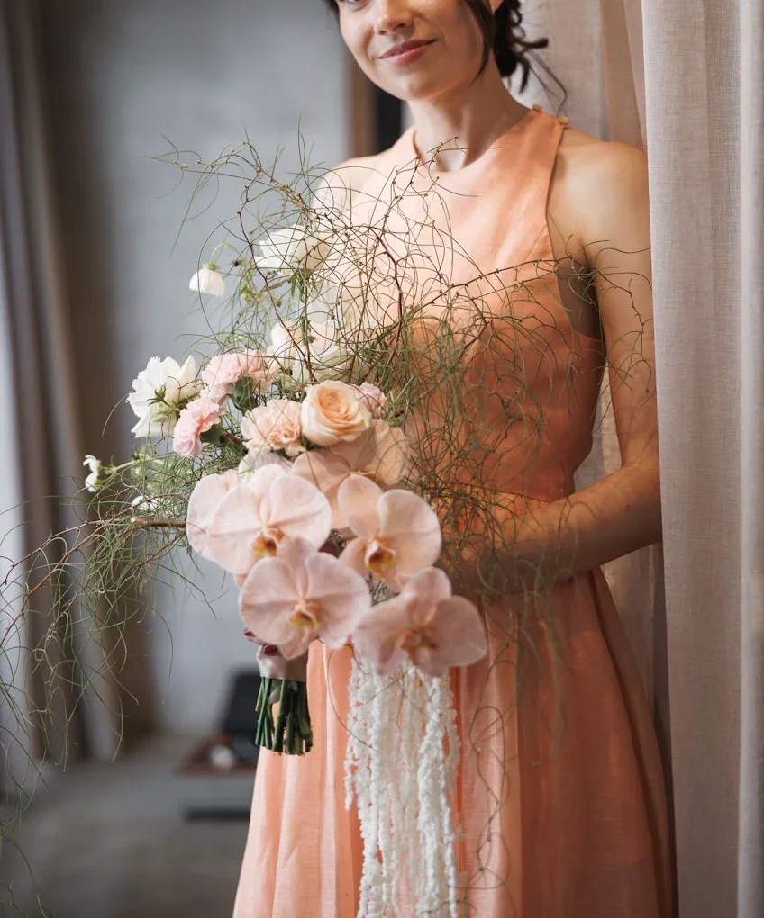 A woman in an orange dress holding a large bouquet of pink and white flowers, including orchids, roses, and other blooms, with green and brown foliage.