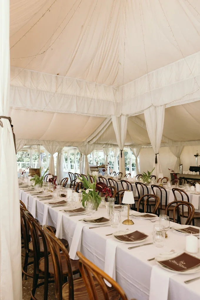 Elegant indoor event tent with white draped ceiling and long dining tables set with white tablecloths, brown plates, glasses, and floral centerpieces.