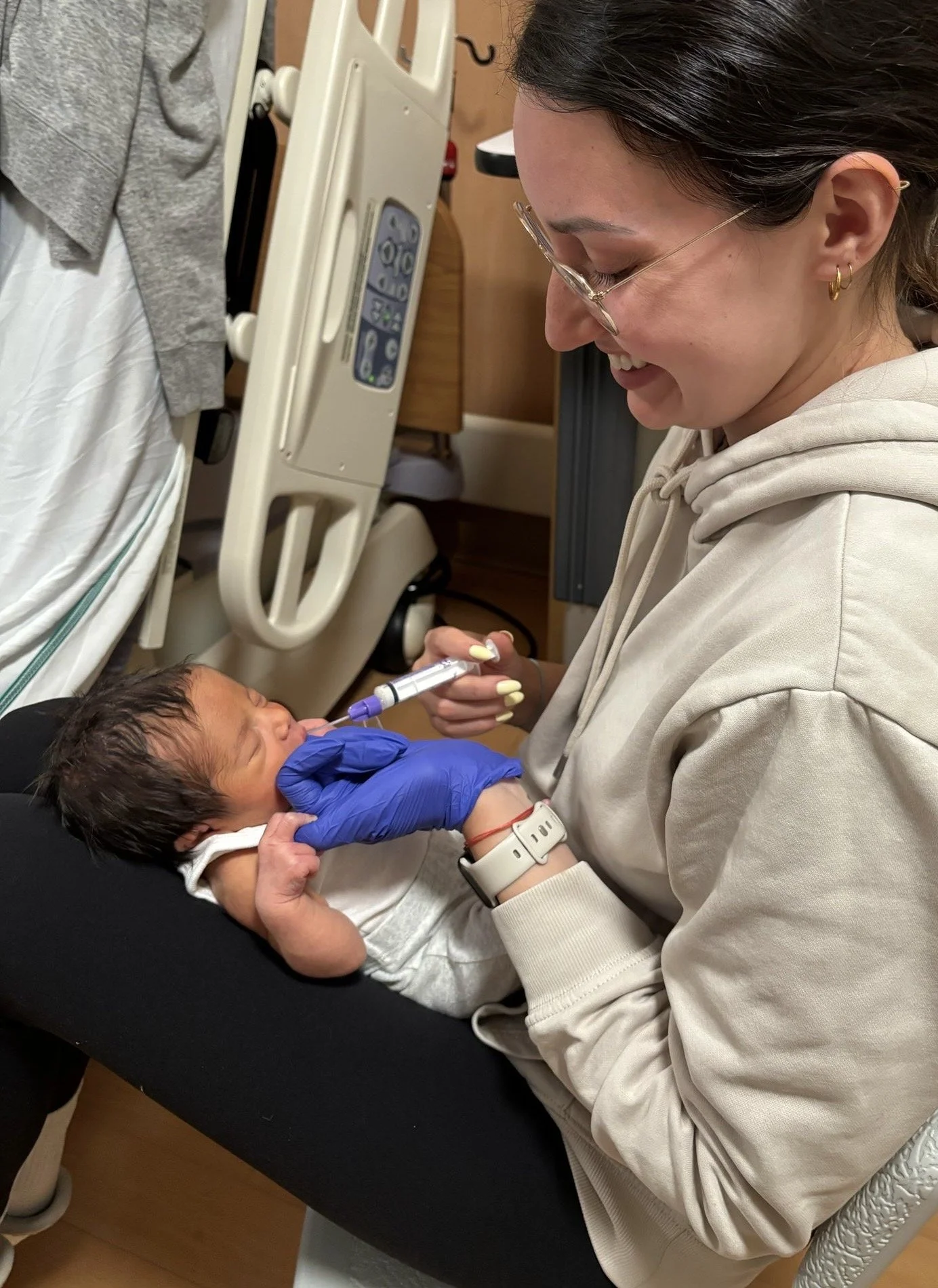 A woman with glasses sitting on a hospital bed, smiling and holding a newborn baby. The baby is receiving a medical injection from a healthcare professional wearing blue gloves, while placenta cloth is visible in the background.