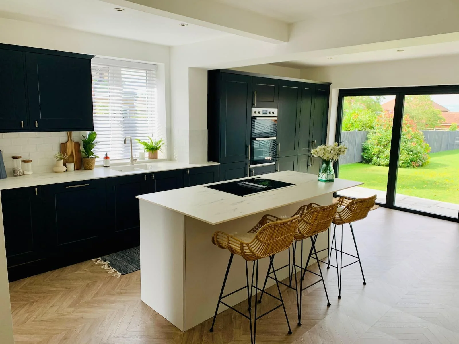 Modern kitchen with black cabinets, white countertops, an island with four wicker bar stools, and large sliding glass doors leading to a backyard.