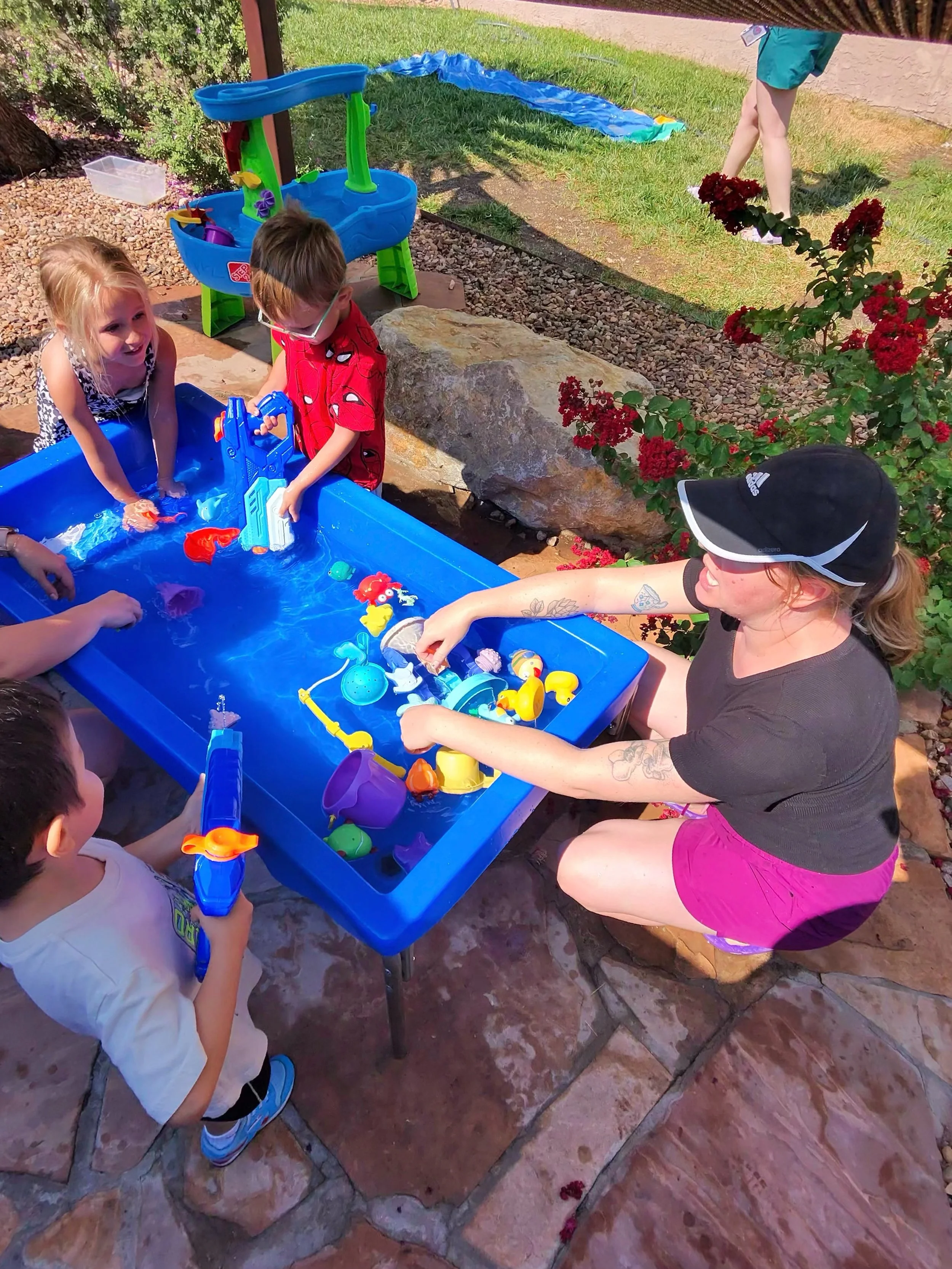 Children playing with water toys in an outdoor patio setting, supervised by an adult woman wearing a black cap and pink shorts.