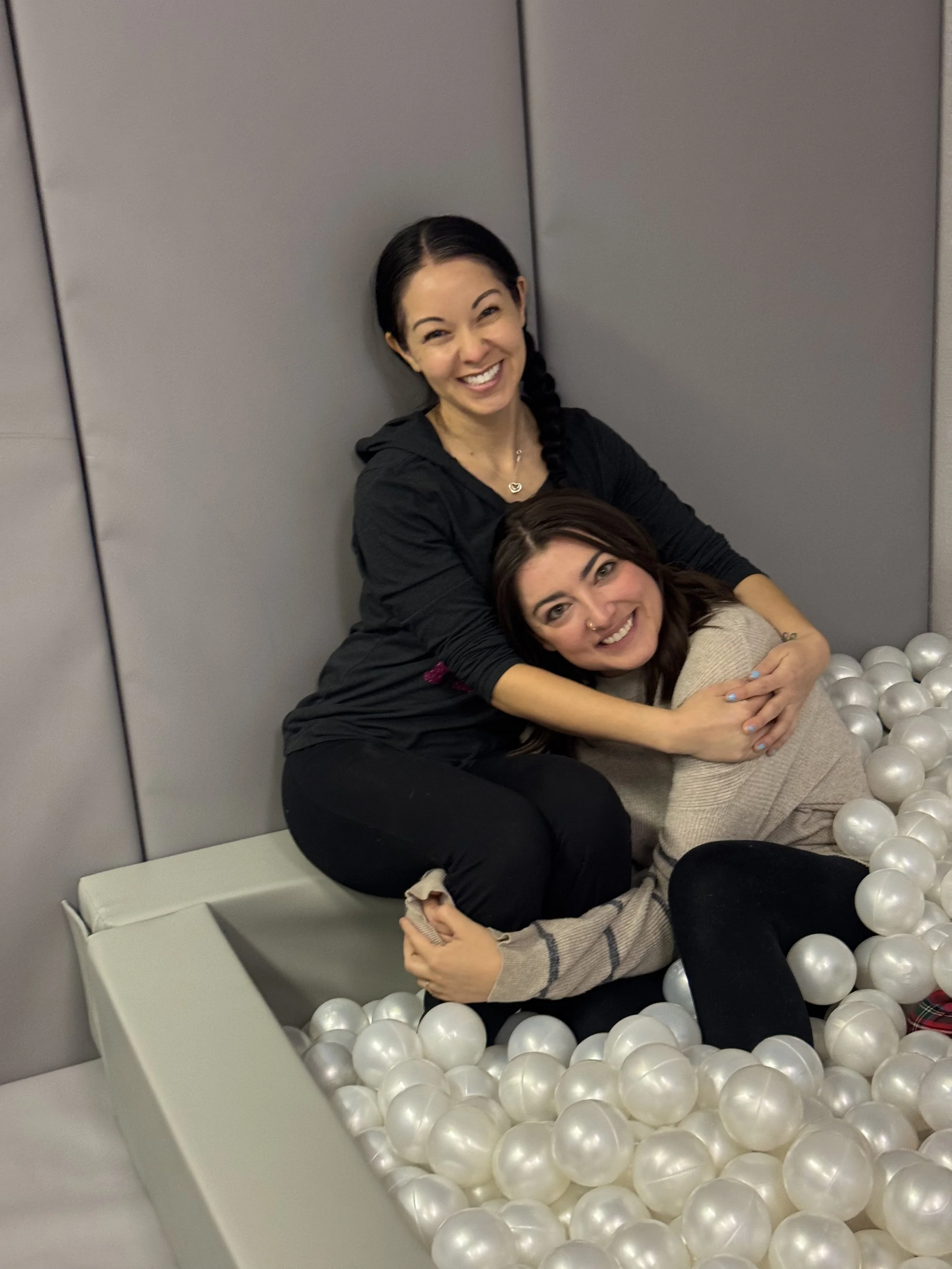Two women sitting and smiling in a ball pit filled with white balls, against gray wall background.
