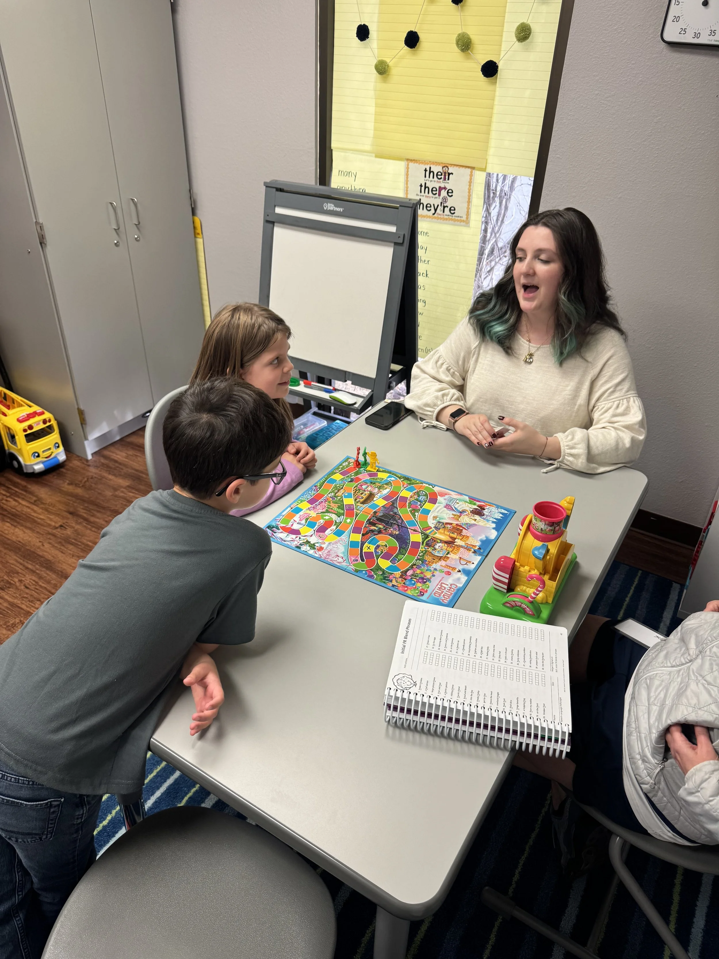 A teacher and two children playing a board game at a table in a classroom.