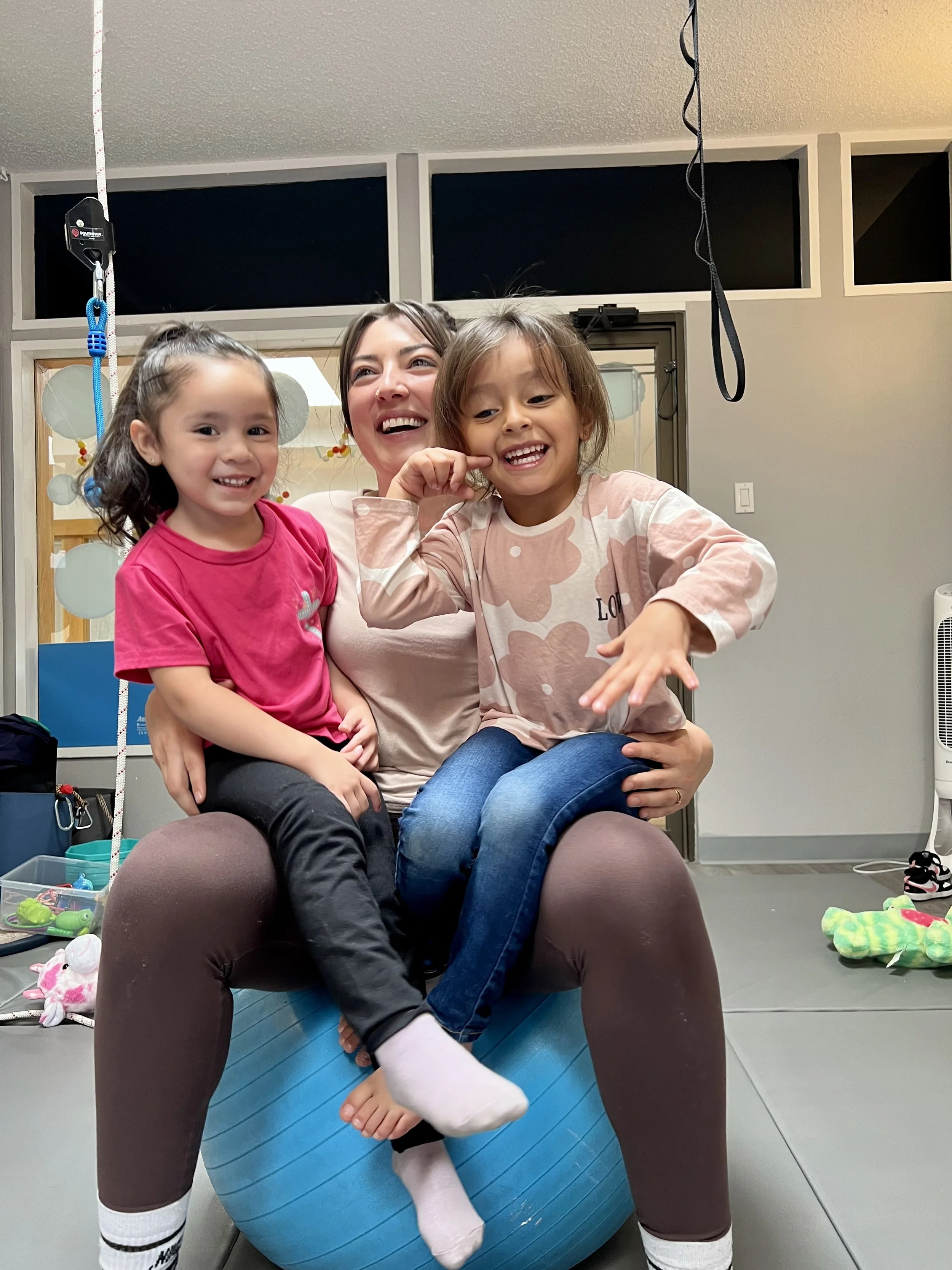 A woman and two young girls sitting on an exercise ball, smiling and laughing in a gym or playroom. The woman is in the middle, with the girls to her sides.