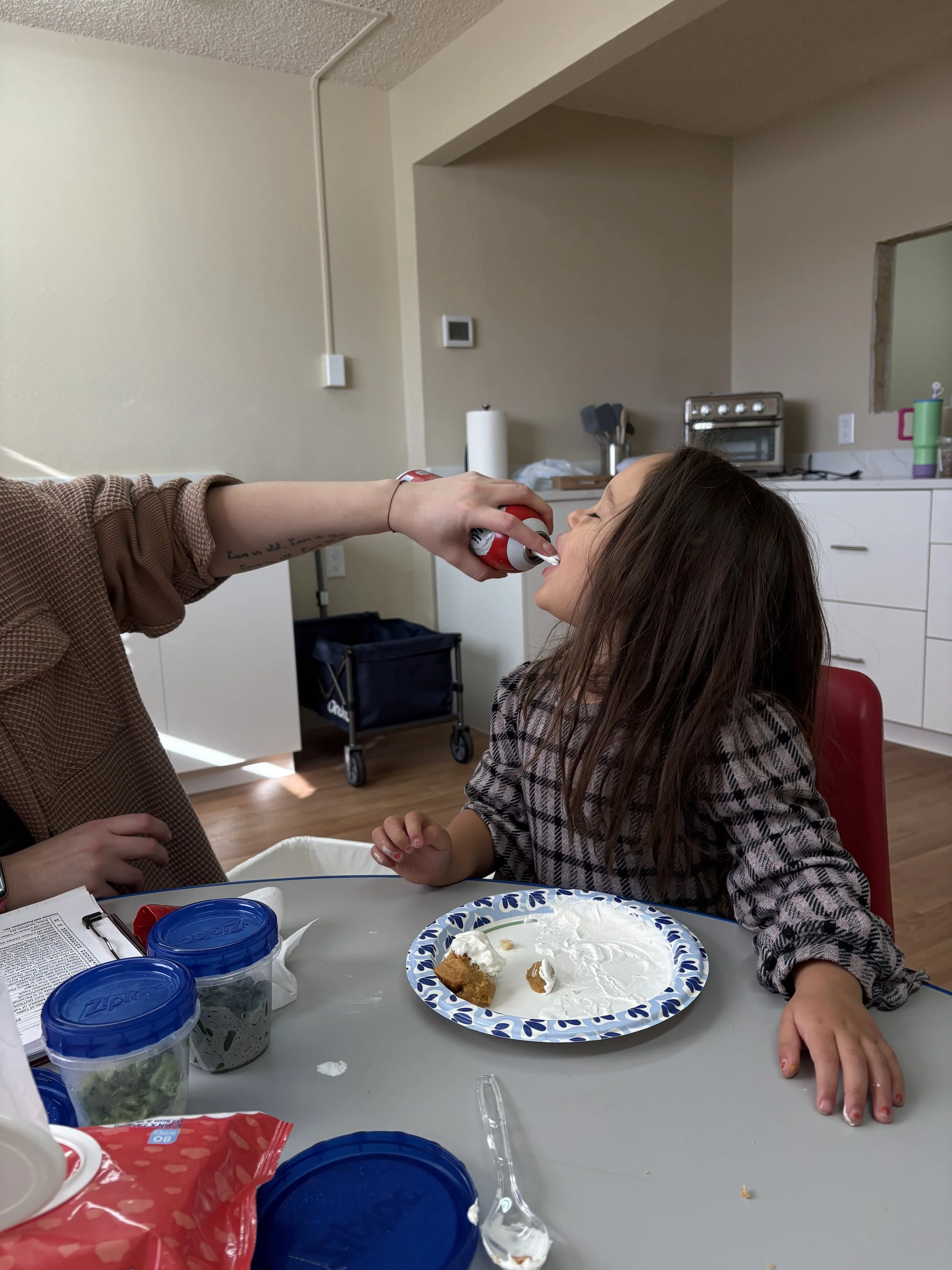 A girl with long brown hair sitting at a table with a paper plate of cake in front of her, as someone sprays her with whipped cream in her open mouth.