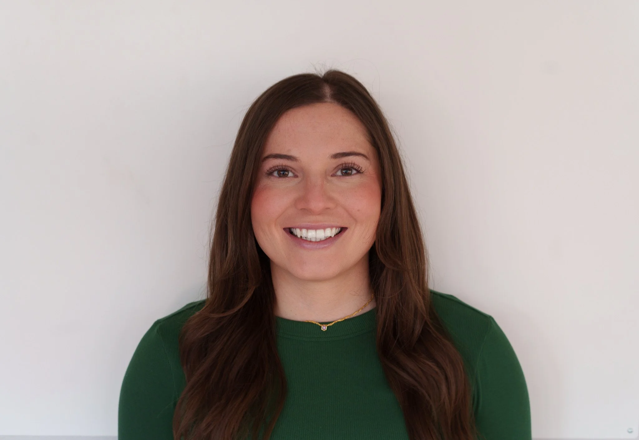 A woman with long brown hair, smiling, wearing a green top and a delicate gold necklace, standing against a plain white background.