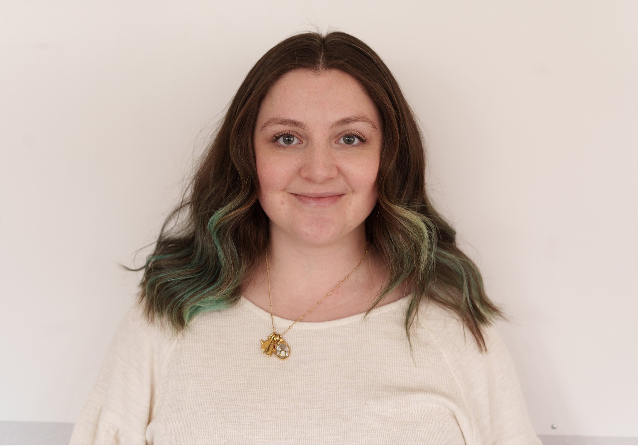 A woman with shoulder-length brown hair with green highlights, wearing a cream-colored top and a gold necklace with charms, smiling against a plain white wall.