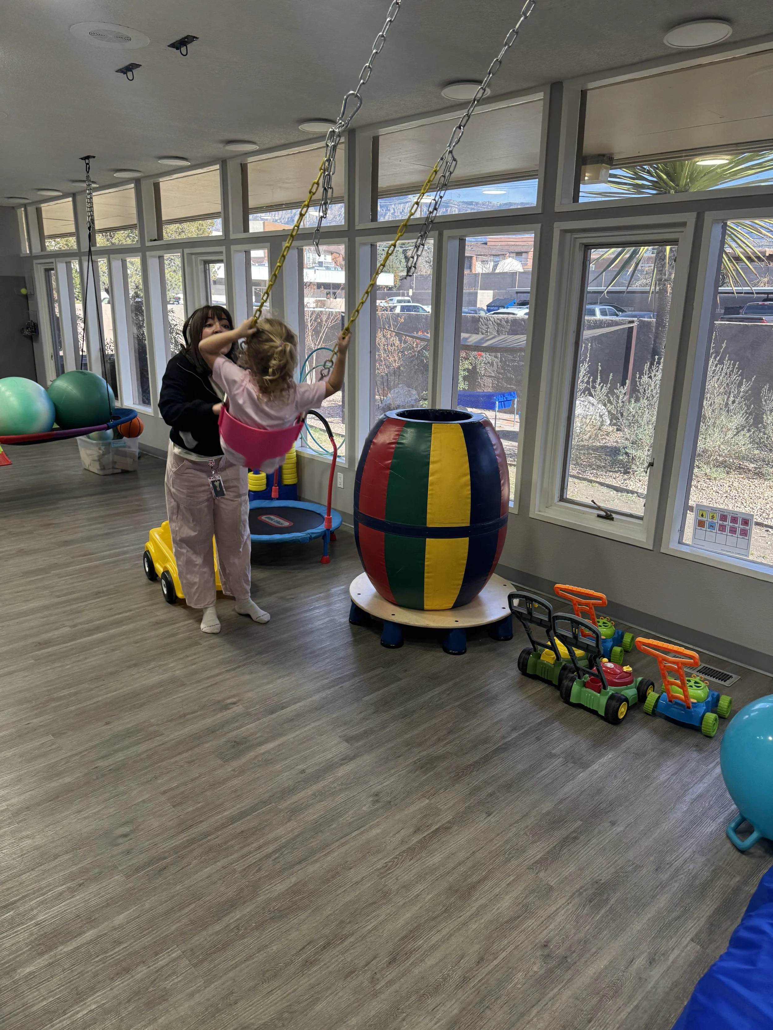 Children playing with a large colorful barrel swing in a playroom with windows, toys, and fitness balls.