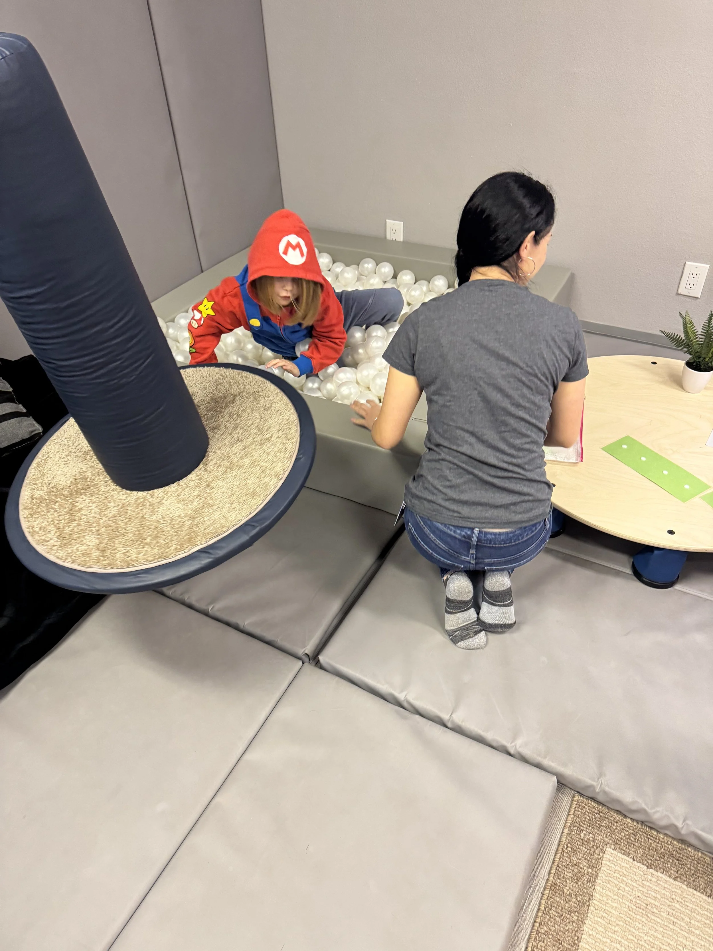 A woman kneeling on gray mats plays with a child in a ball pit filled with white plastic balls. The child wears a red hoodie with a Mario logo. There is a round table with a small plant and a green paper on it, and part of a black punching bag is visible.