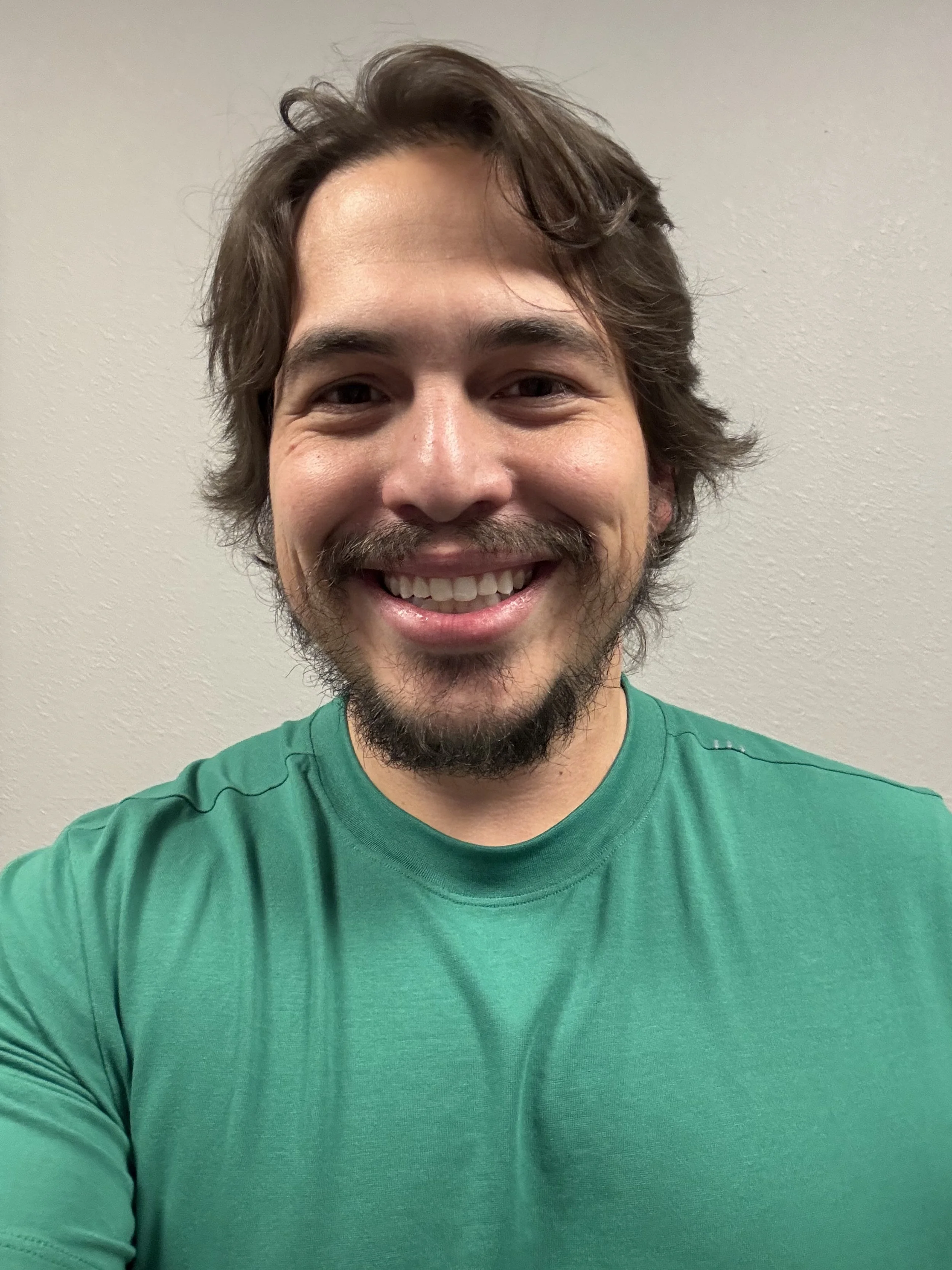A smiling man with brown hair, a beard, and mustache taking a selfie against a plain beige wall, wearing a green shirt.