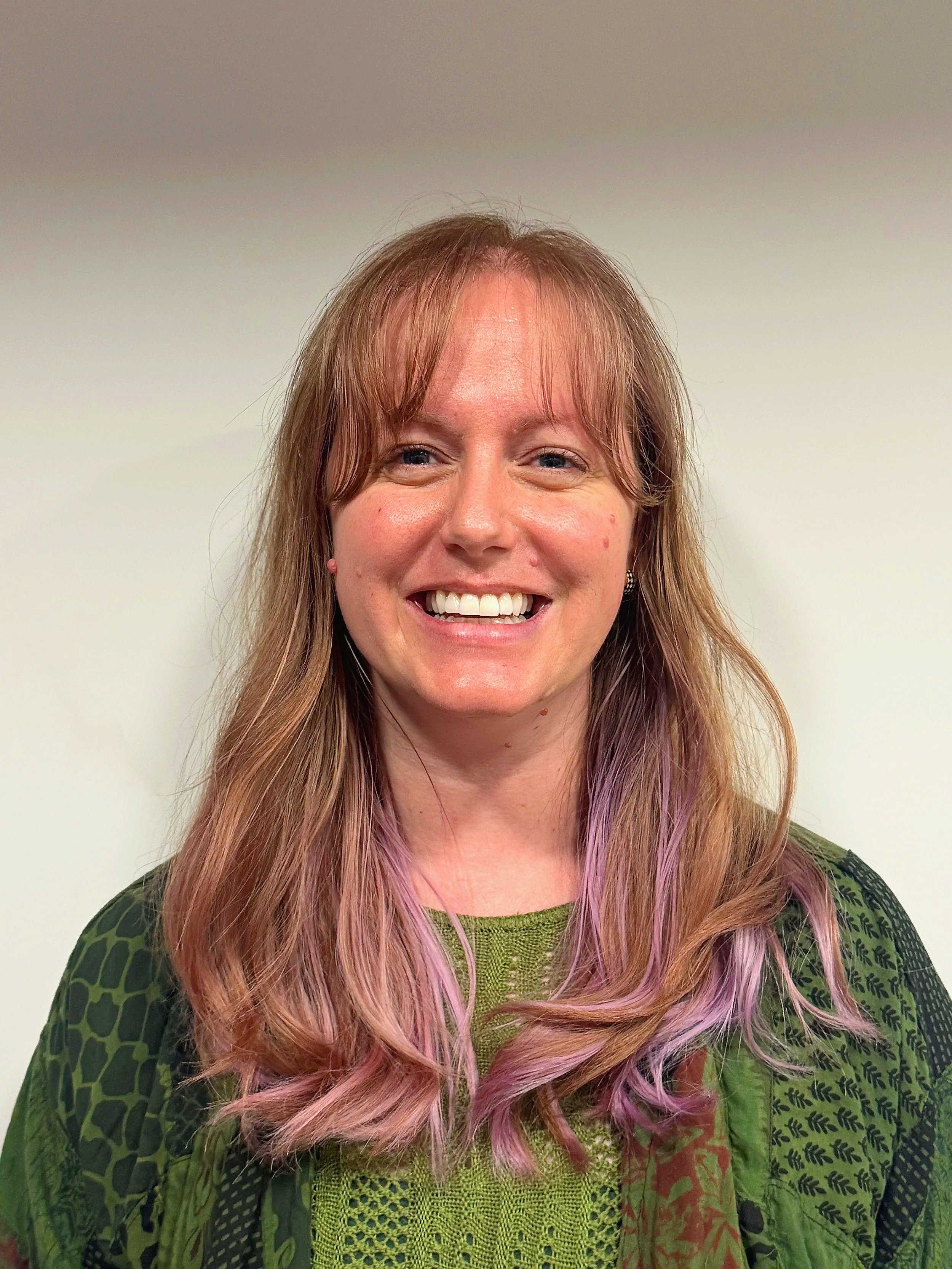 A woman with light skin, reddish-brown hair with pink highlights, smiling against a plain white wall background.