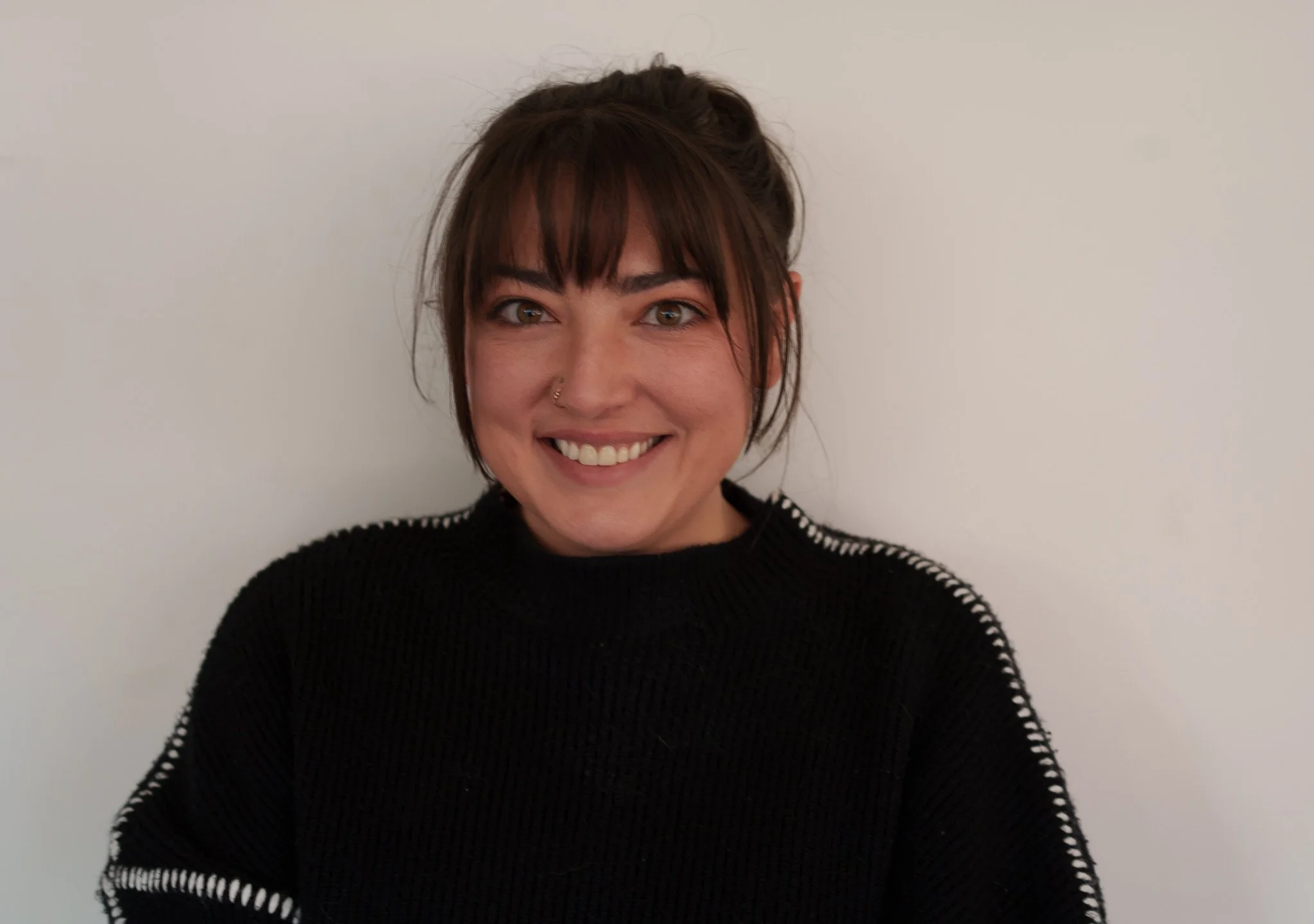 A woman with short brown hair and bangs, smiling, wearing a black sweater with white stitching on the sleeves, standing against a plain white wall.