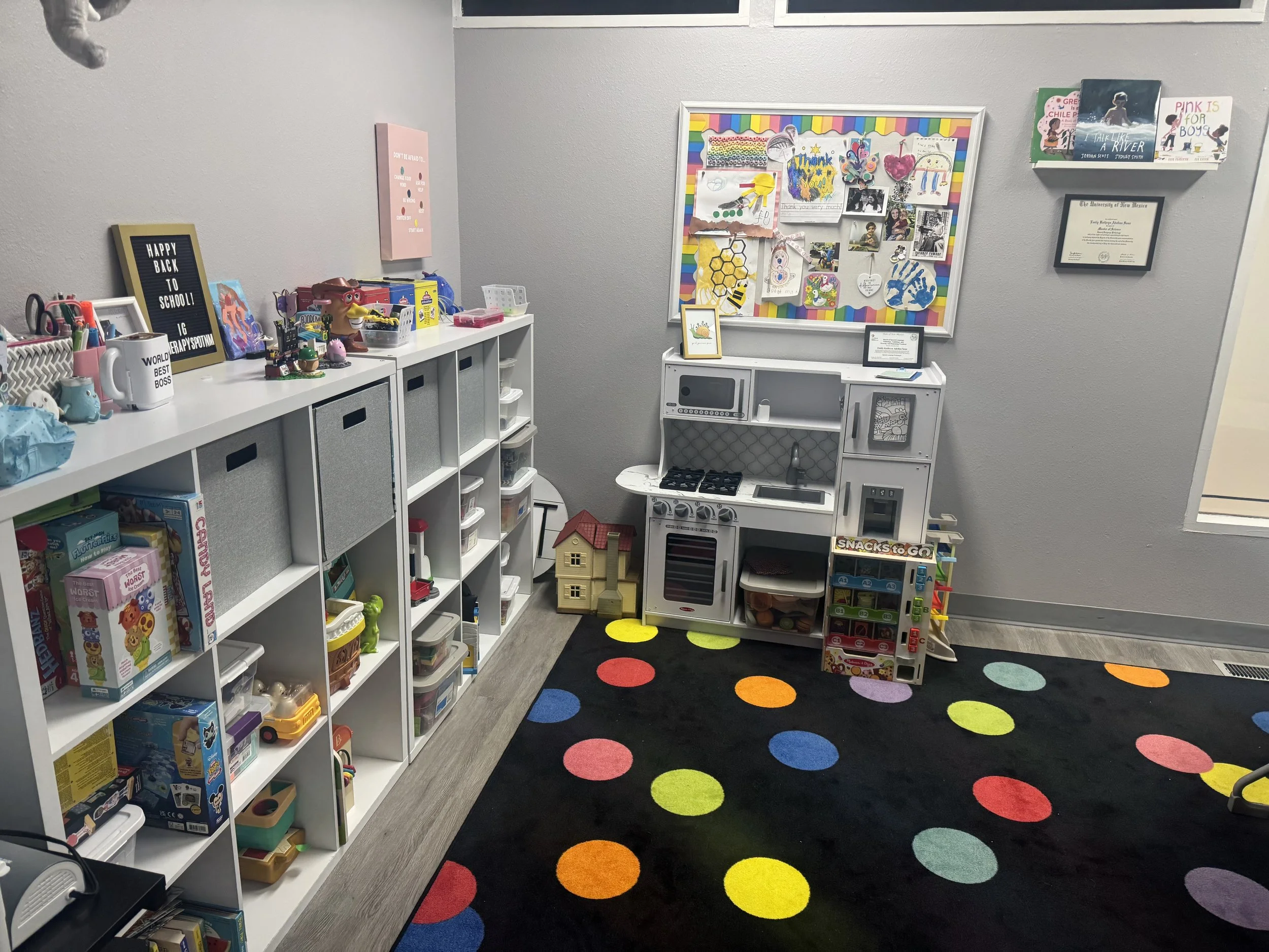 A children's playroom with a white storage unit filled with toys and books, a colorful play kitchen, a black rug with multicolored dots, and framed artwork on the gray wall.