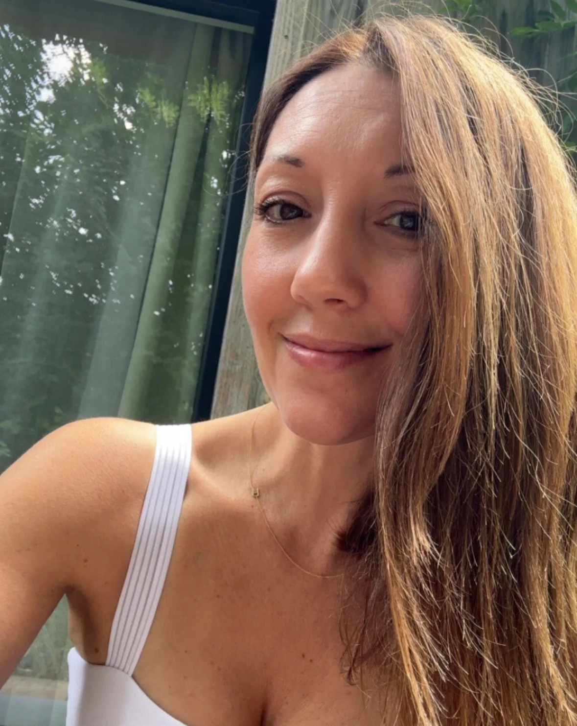 Close-up of Rachel with long reddish-brown hair, smiling, wearing a white strap top and a delicate gold necklace, with a window and greenery in the background.