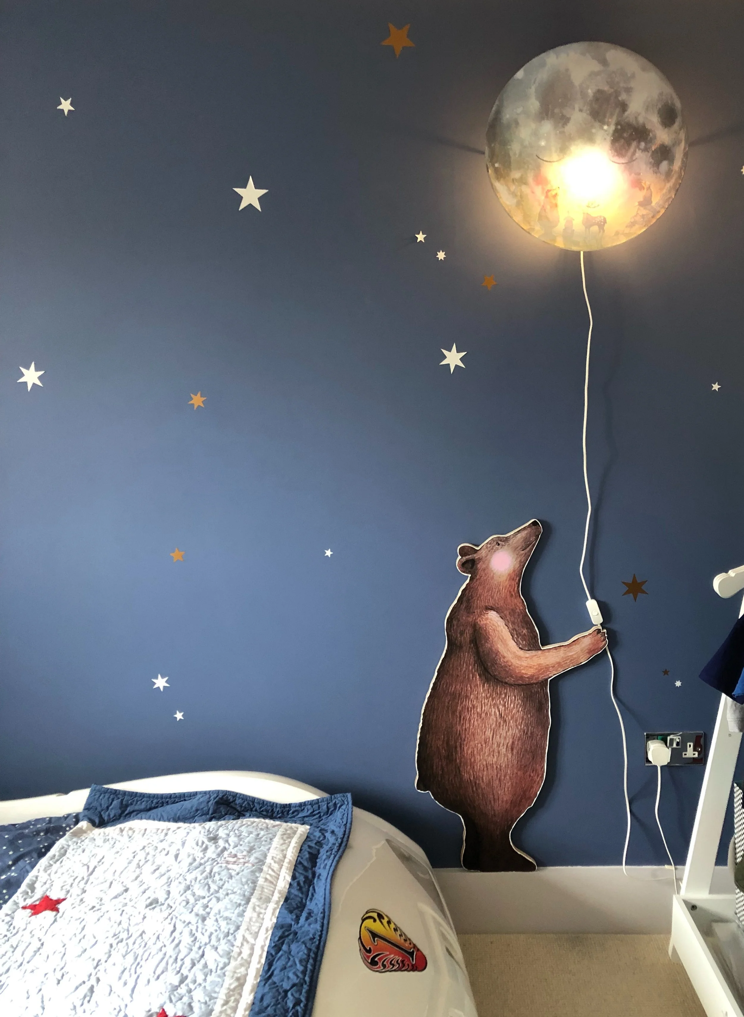 A navy blue wall decorated with star stickers and a moon-shaped light fixture. A wall decal of a bear holding a balloon string is attached to the wall. Part of a bed with a blue and white quilt and a racing car sticker is visible in the foreground.