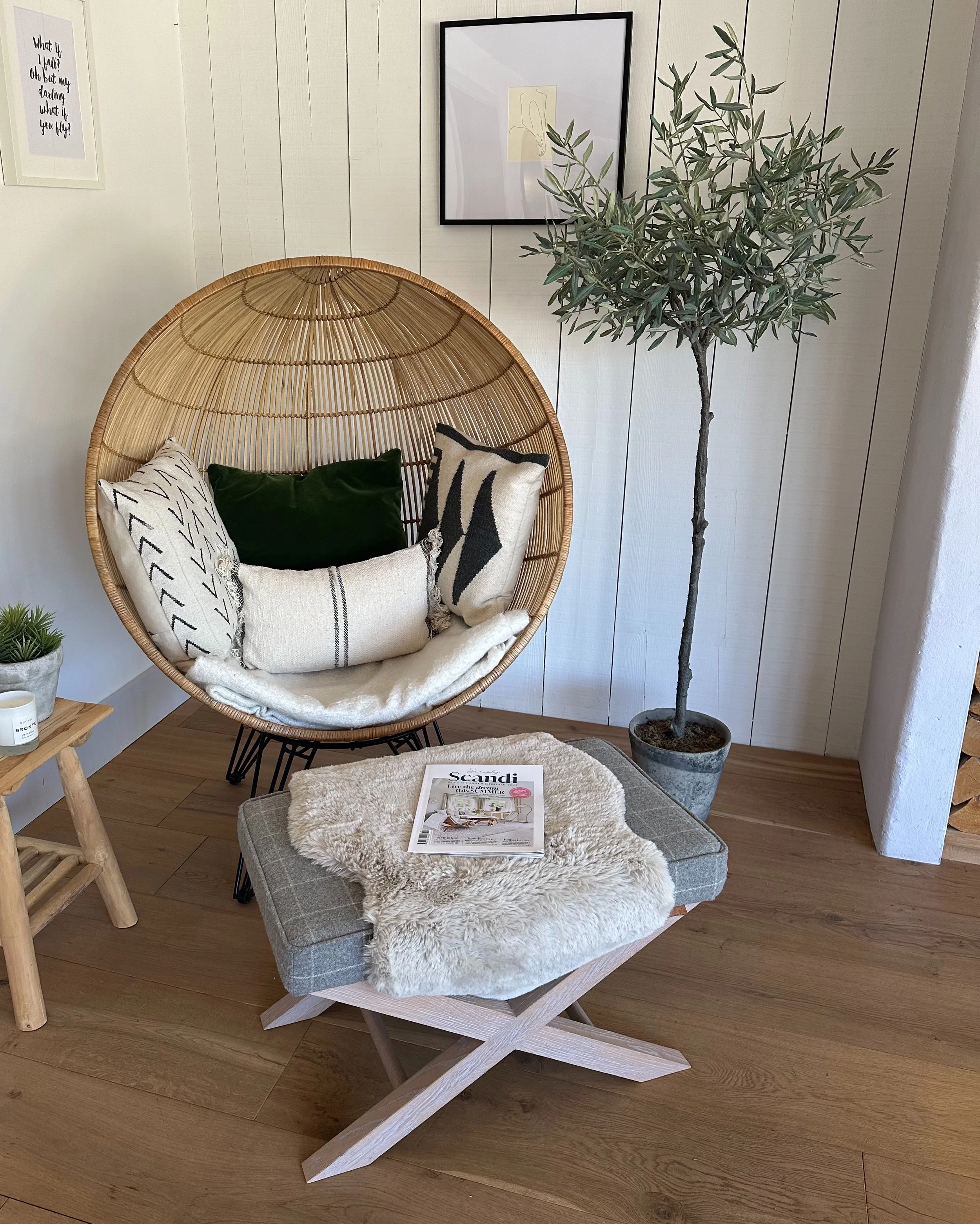 Cozy corner with a hanging papasan chair with pillows, a small wooden side table with a plant and candle, a gray ottoman with a soft blanket and magazine, and a tall potted olive tree in a vase, with framed art and quotes on white shiplap walls.