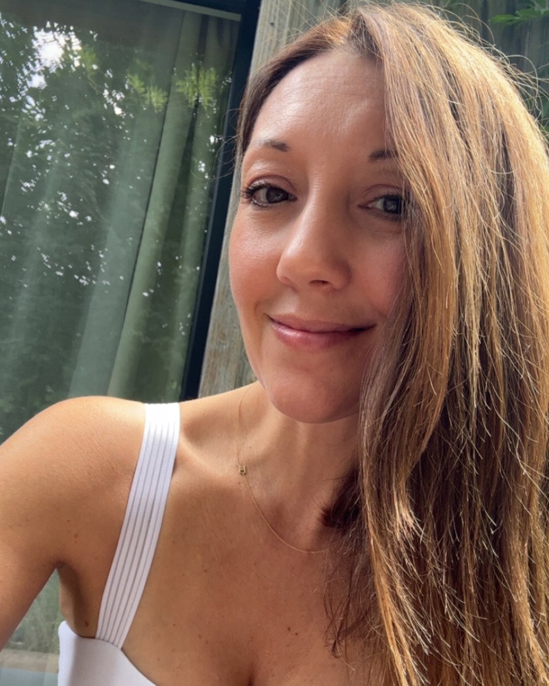 A woman with long, reddish-brown hair, wearing a white top, smiling outdoors with trees and glass window in the background.