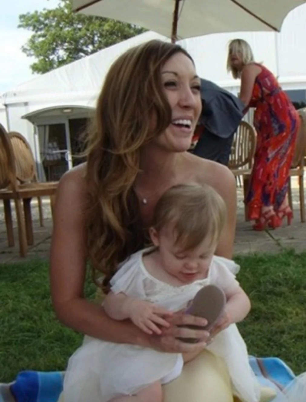Woman sitting outdoors with a young child, both laughing and looking at a smartphone. In the background, there is a woman in a red dress and a large white umbrella.