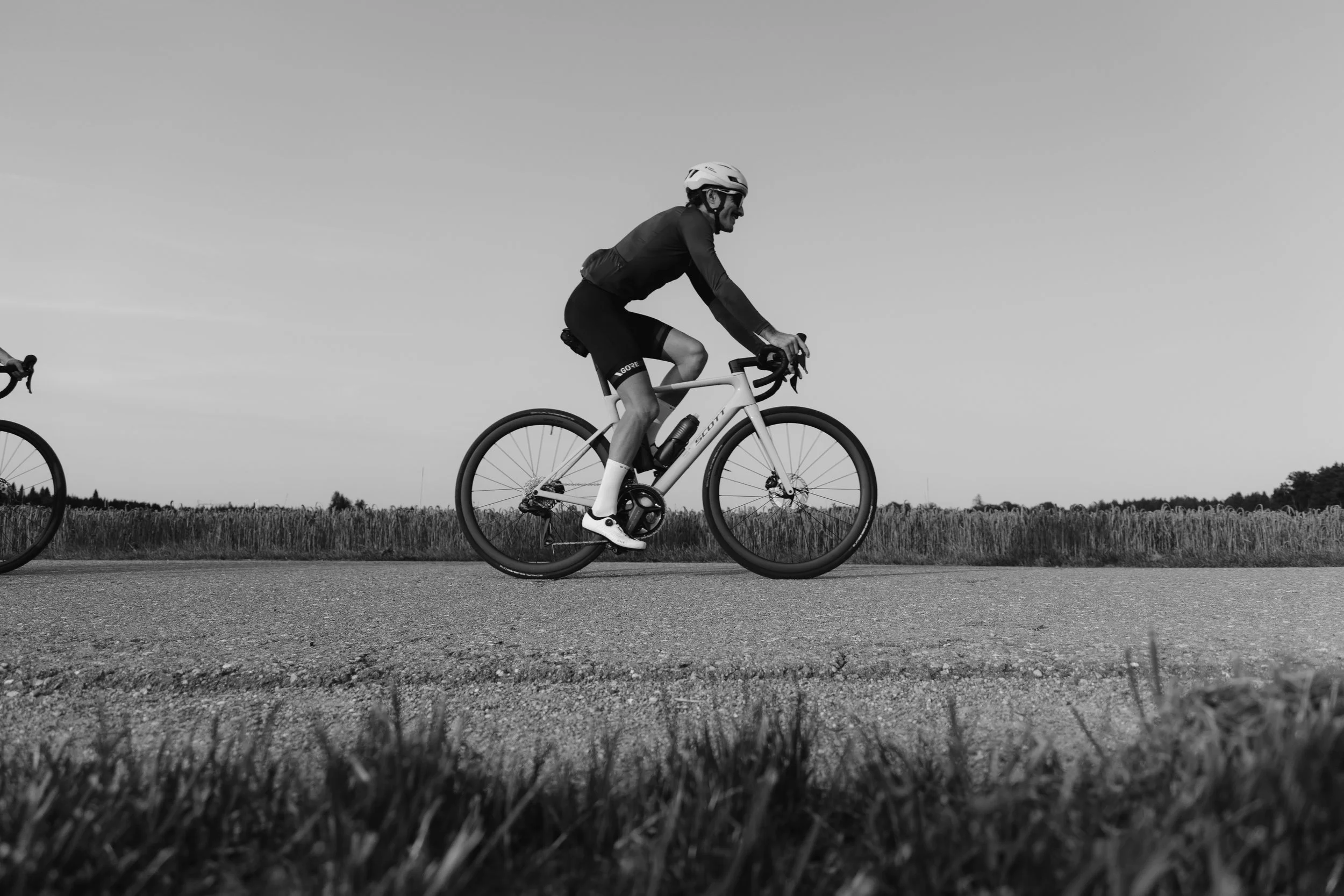 Mann fährt Fahrrad auf einer Landstraße, trägt Helm und Sportbekleidung, im Hintergrund Felder, schwarz-weiße Aufnahme.