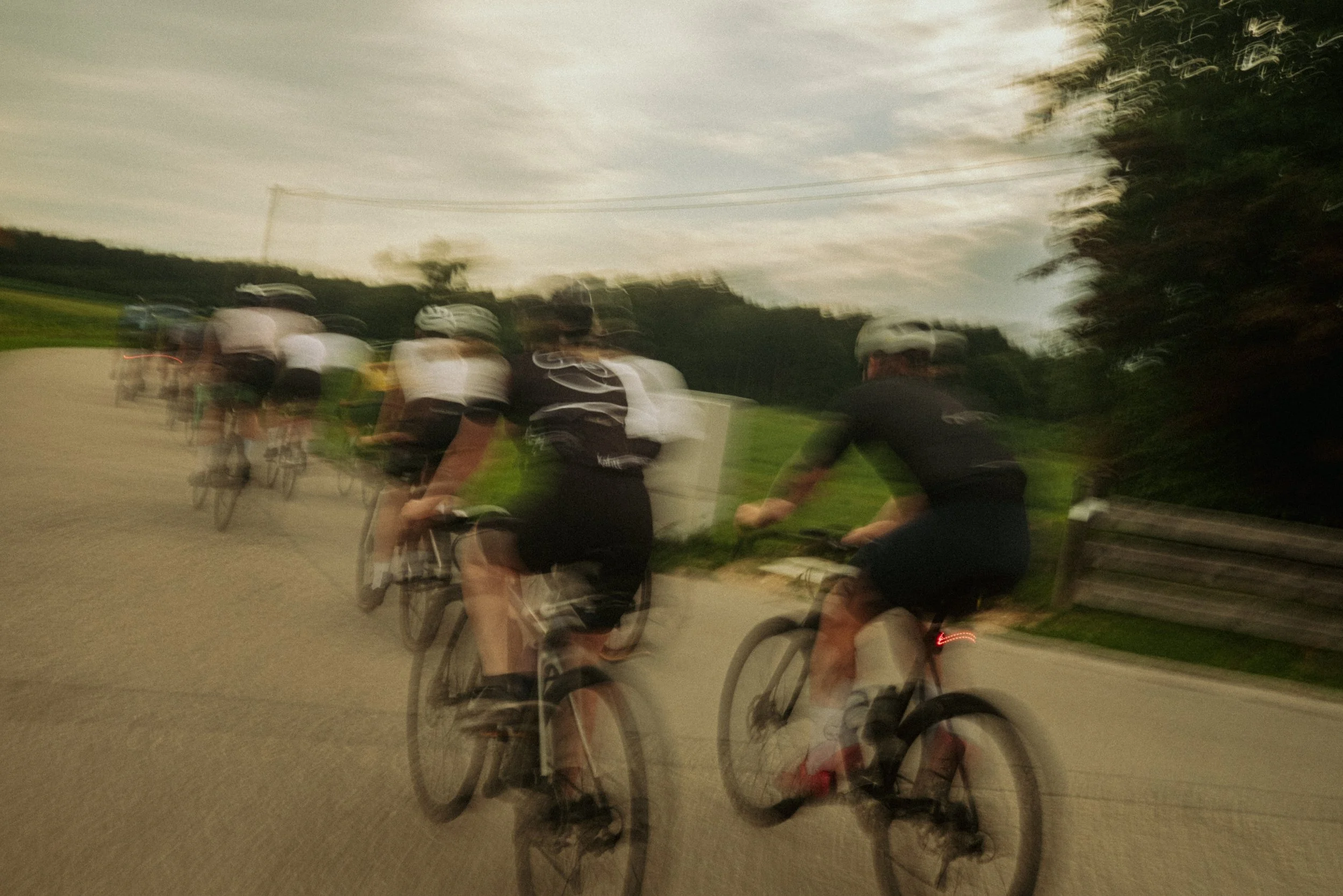 Mehrere Fahrradfahrer in Bewegung, unscharf aufgenommen, unterwegs auf einer Straße im Freien bei Dämmerung.