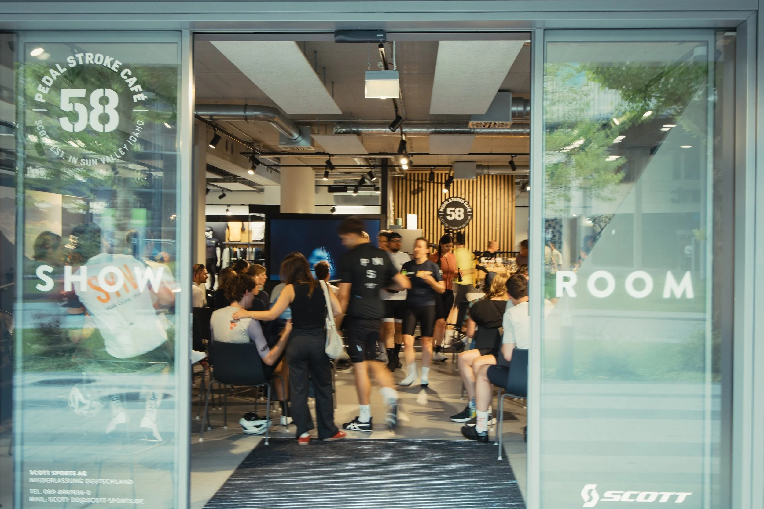 Eingang zum Scott Sports Laden mit Blick ins Geschäft, in dem Menschen an einem Vortrag oder Meeting teilnehmen.