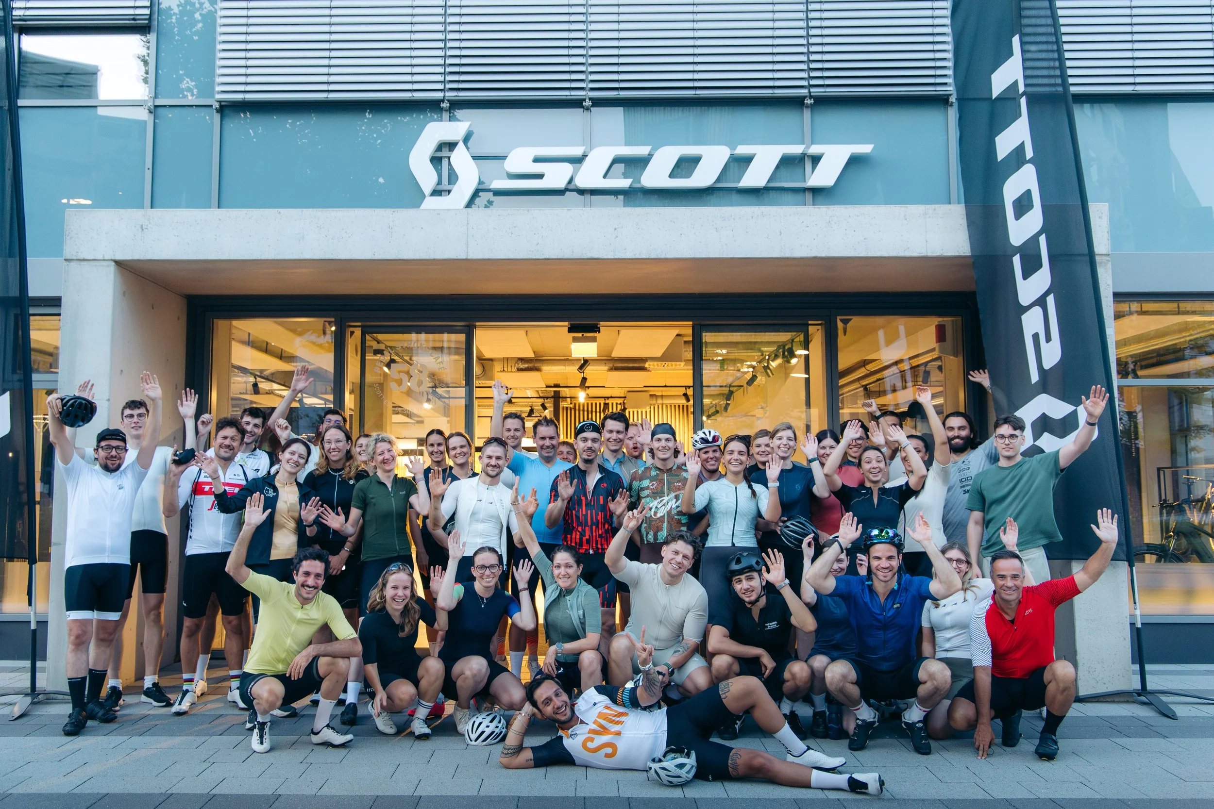 Gruppe von Menschen vor einem SCOTT Geschäft, die jubeln und die Hände heben, einige tragen Fahrradhelme, bei einem sportlichen Veranstaltungs- oder Team-Event.