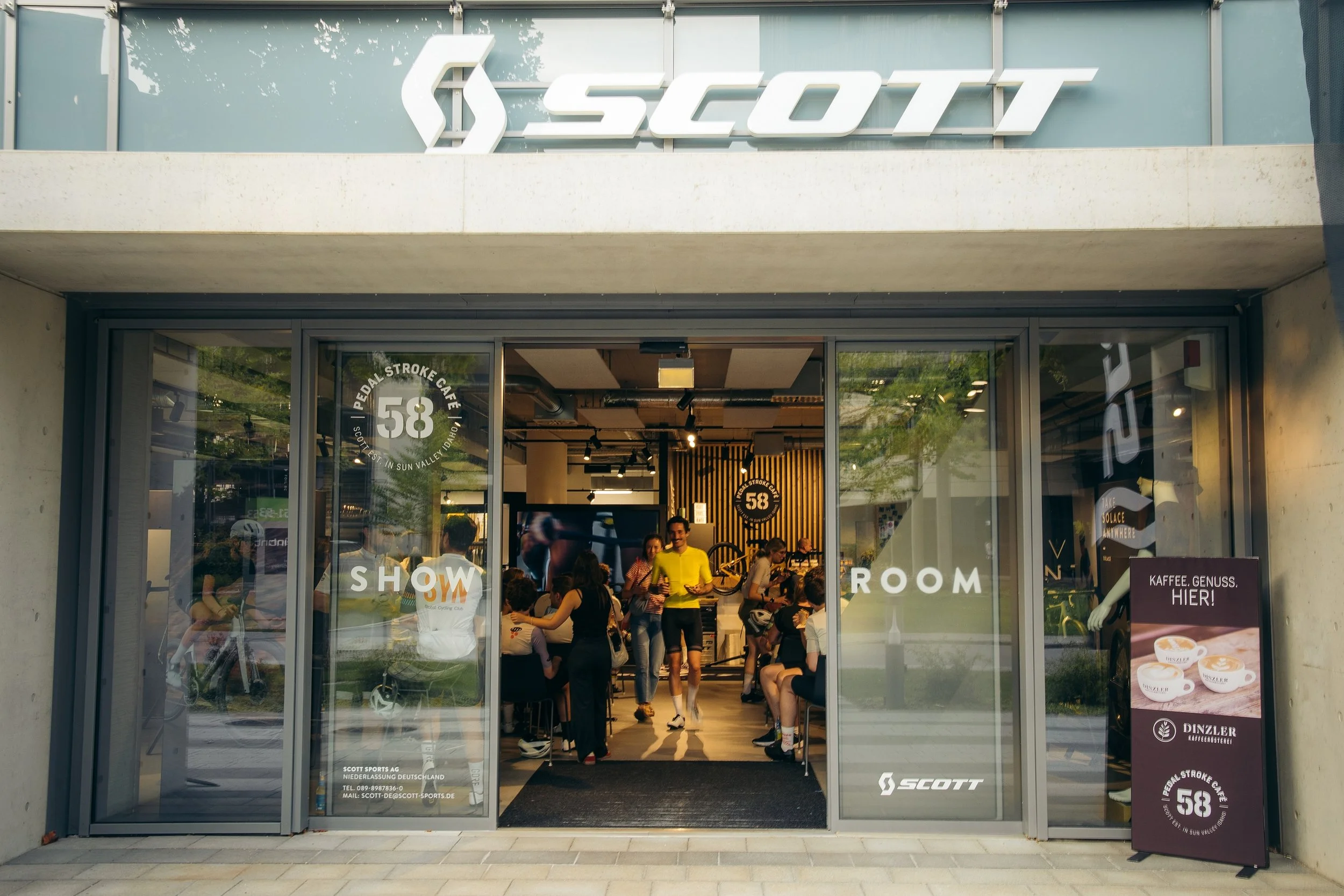 Das Bild zeigt den Eingangsbereich eines Cafés oder Geschäfts namens Scott mit großen Glasschiebewänden. Innen befinden sich mehrere Menschen, einige sitzen, während andere stehen und sich unterhalten. Es gibt eine Fahrradmarke Scott im Vordergrund, 